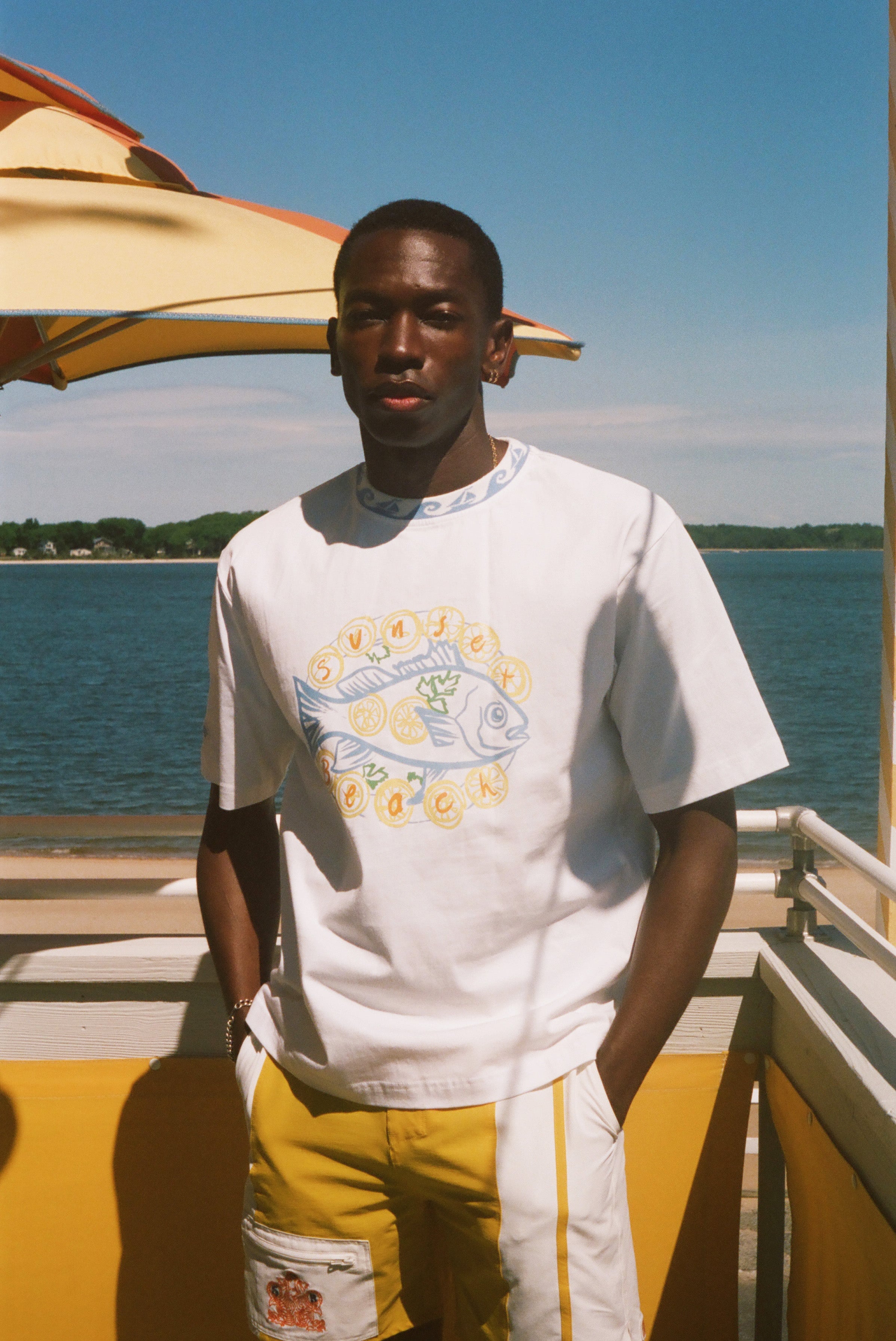 A man wearing a white graphic t-shirt with a blue and yellow fish design and yellow shorts, standing on a sunny boat deck with water and a blue sky in the background.