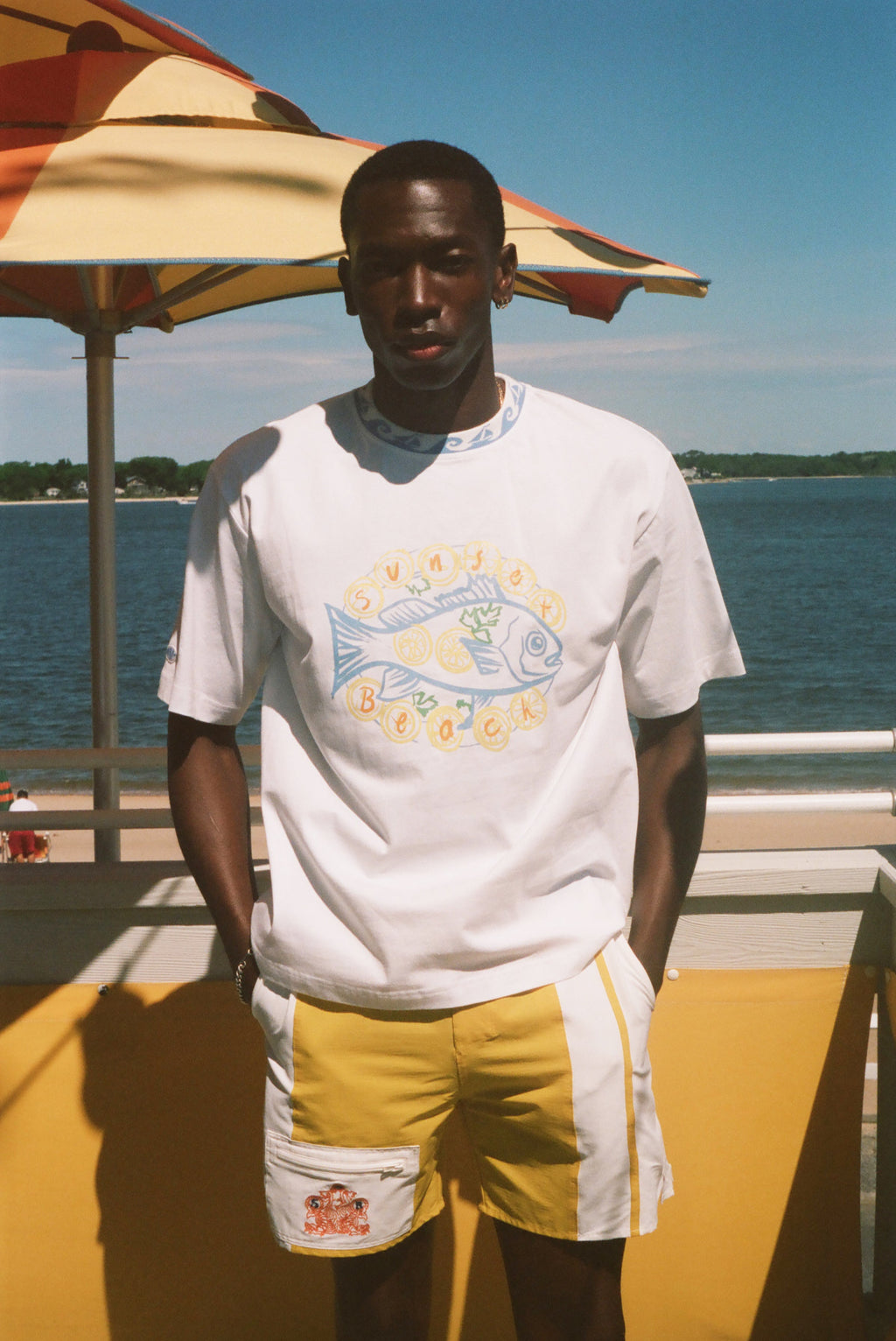 A man standing outdoors by the water wearing a white graphic T-shirt with a large fish design and yellow shorts, with a beach umbrella and blue sky in the background.