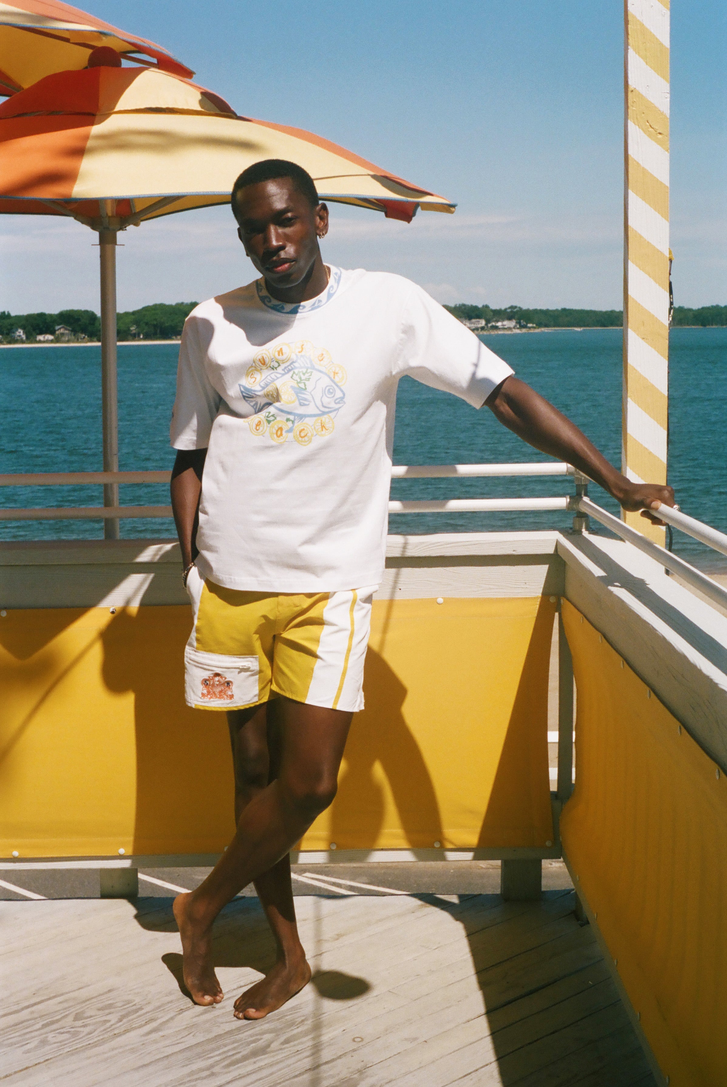 A man stands on a sunny outdoor deck by the water wearing a white graphic T-shirt and yellow shorts with white side stripes. An orange-and-white striped umbrella, yellow railings, and blue water are visible in the background.