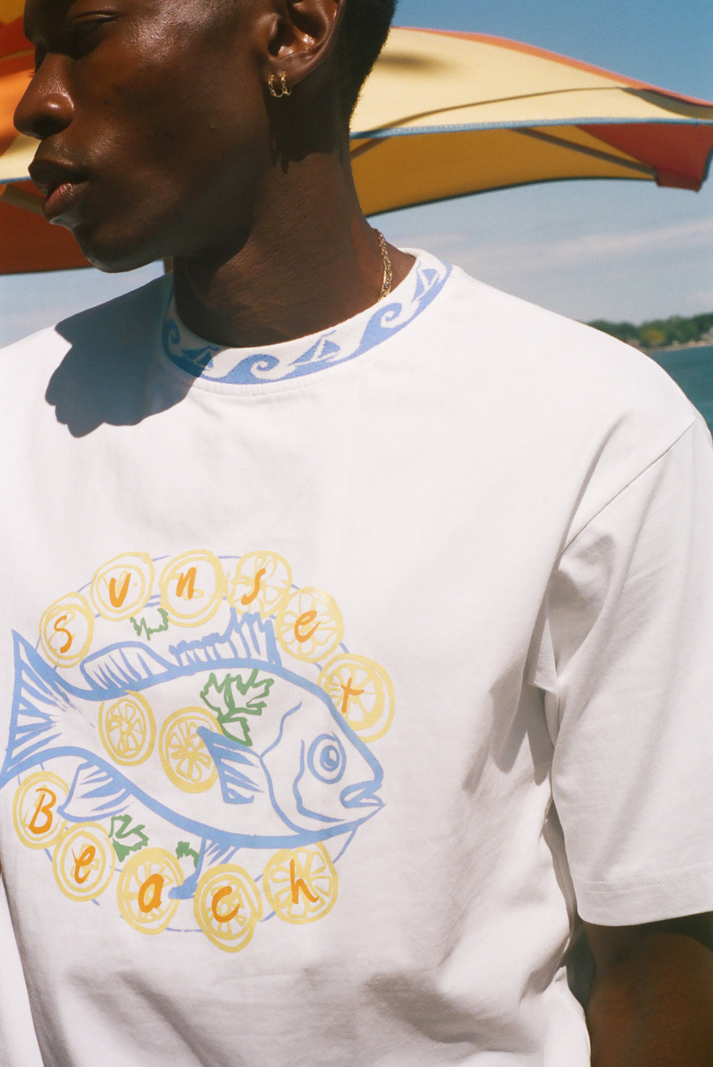 A close-up of a person wearing a white graphic t-shirt with a blue and yellow 'Sunset Beach' fish design, set against a sunny outdoor background with a beach umbrella.