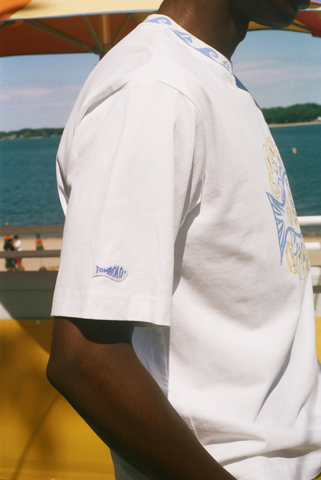 A close-up side view of a person wearing a white short-sleeve t-shirt with a blue and yellow graphic on the back and a small 'Tombolo' logo on the sleeve, set against a background of water and a yellow structure.