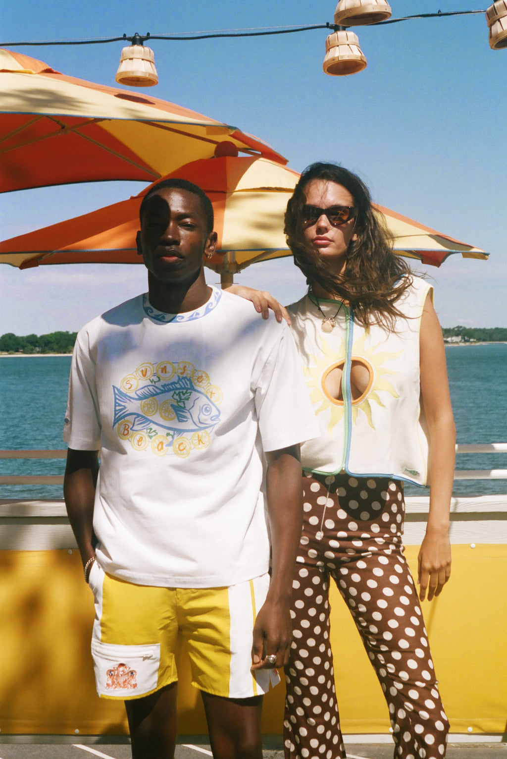 Two models standing outdoors by the water under a red and yellow striped beach umbrella. The model on the left wears a white graphic t-shirt with a blue fish and yellow text, yellow shorts, and a white pocket.