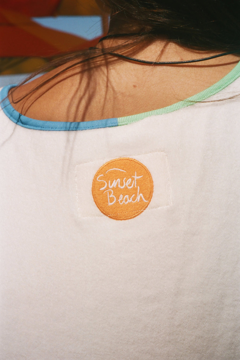 Close-up of the back of a white shirt worn by a person, featuring a circular orange patch with 'Sunset Beach' embroidered in white script. 