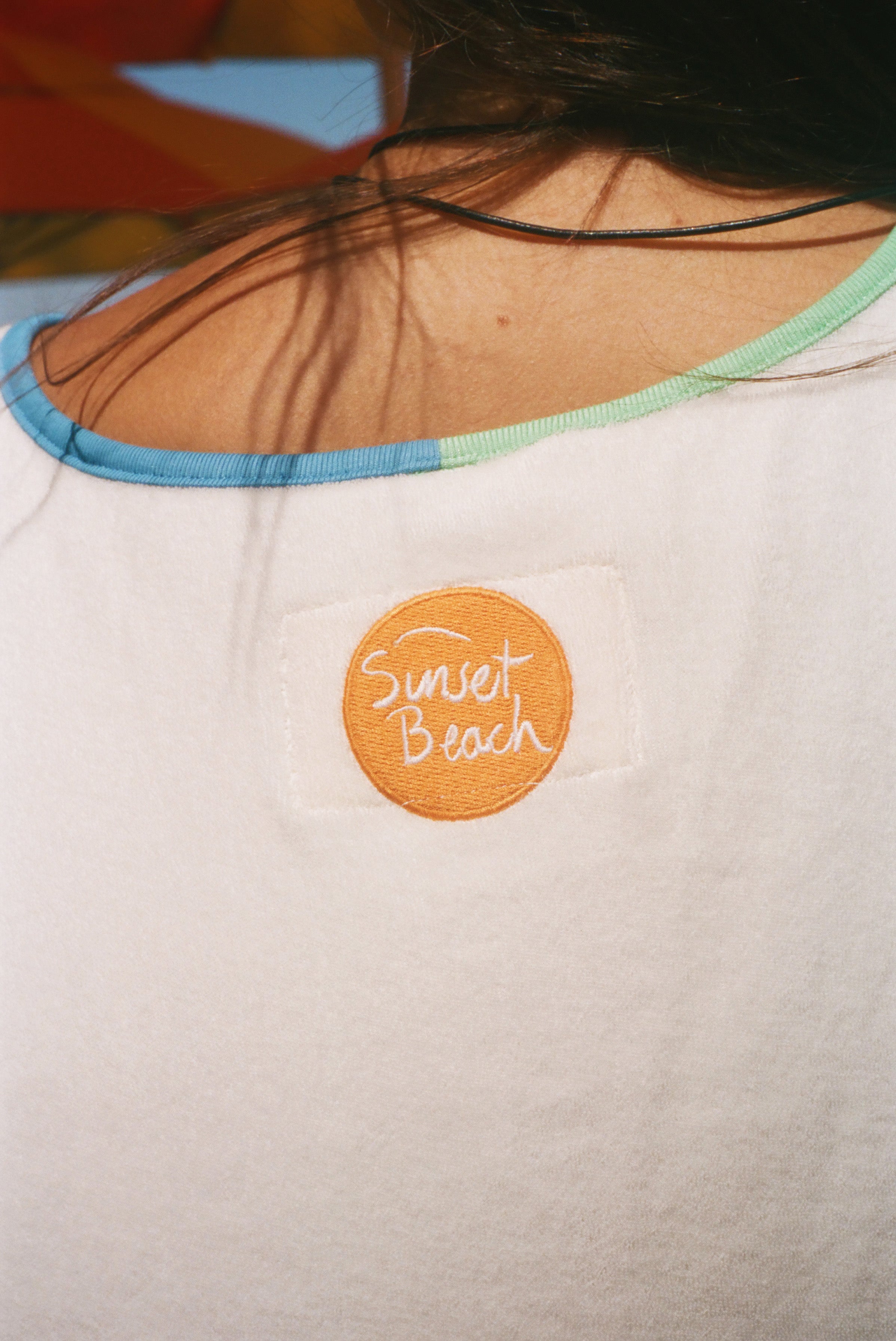 Close-up of the back of a white shirt worn by a person, featuring a circular orange patch with 'Sunset Beach' embroidered in white script. 