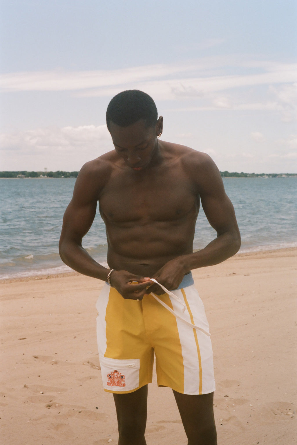 A shirtless man standing on a sandy beach, looking down while adjusting yellow and white swim trunks with a drawstring. The background shows the ocean and a pale sky.