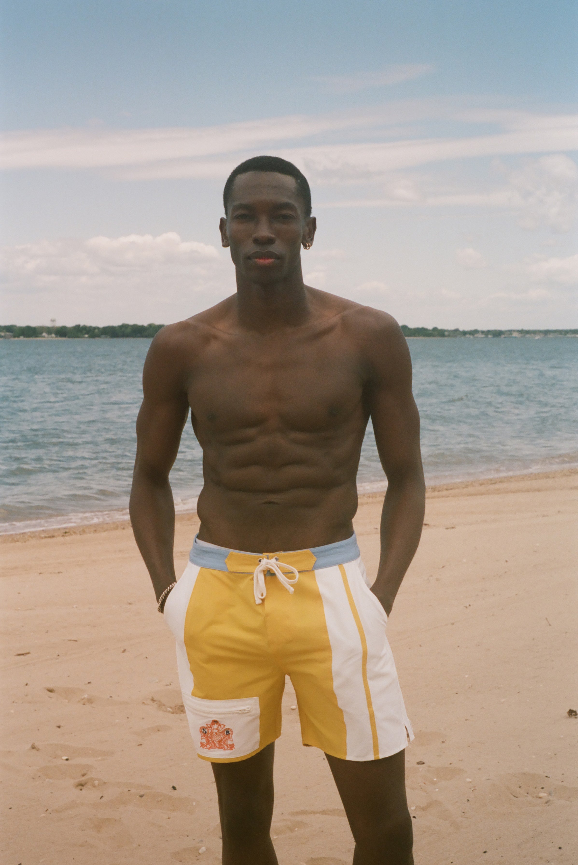 A shirtless man standing on a sandy beach wearing yellow and white swim trunks with a light blue waistband, with the ocean and sky behind him.
