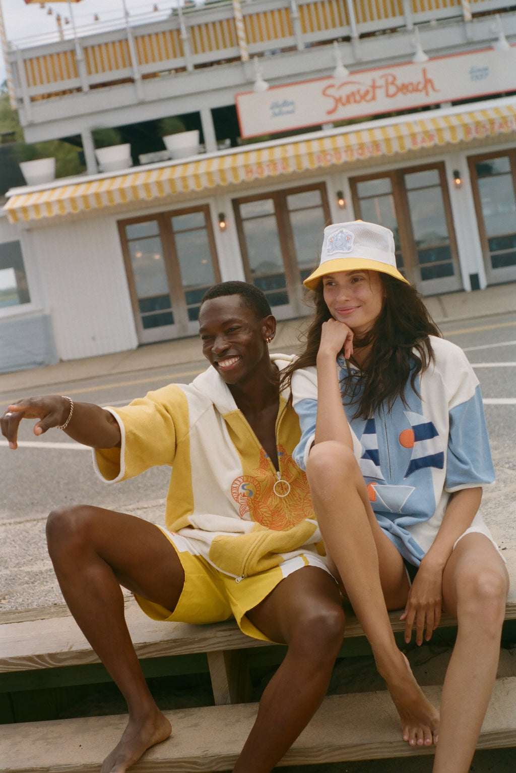 Two models sitting on wooden planks in front of a beachfront restaurant, wearing colorful summer outfits. One model wears a yellow and white short-sleeve shirt and matching shorts, while the other wears a white, blue, and orange graphic t-shirt with a yellow bucket hat.