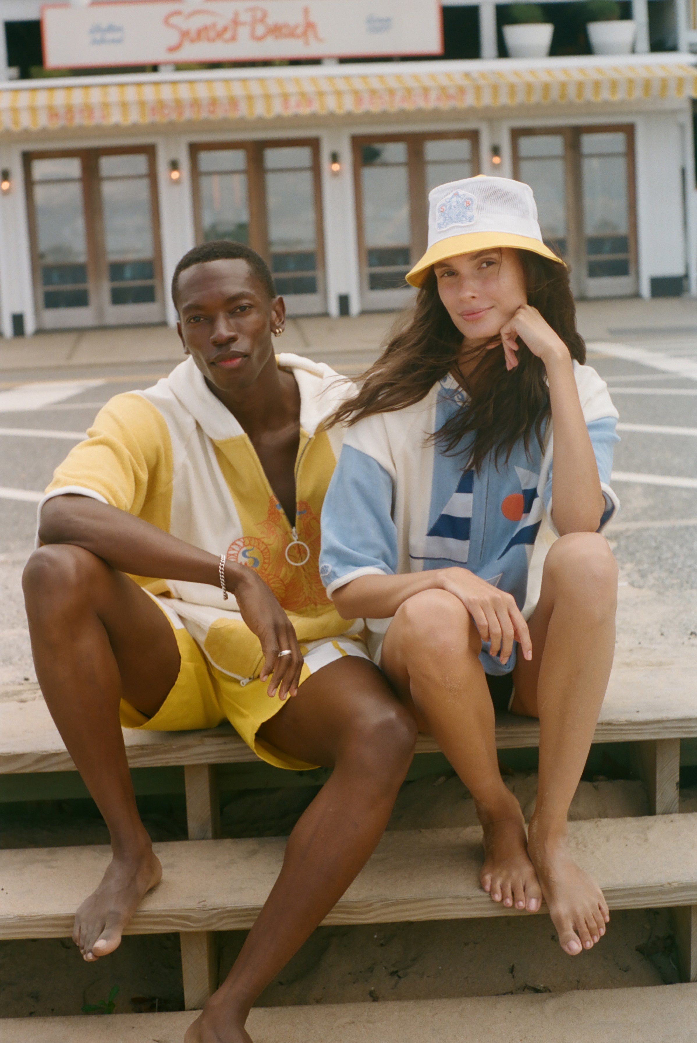 Two models sitting on concrete steps outdoors, wearing coordinated beach-style outfits. The person on the left wears a yellow and white short-sleeve top and matching shorts.
