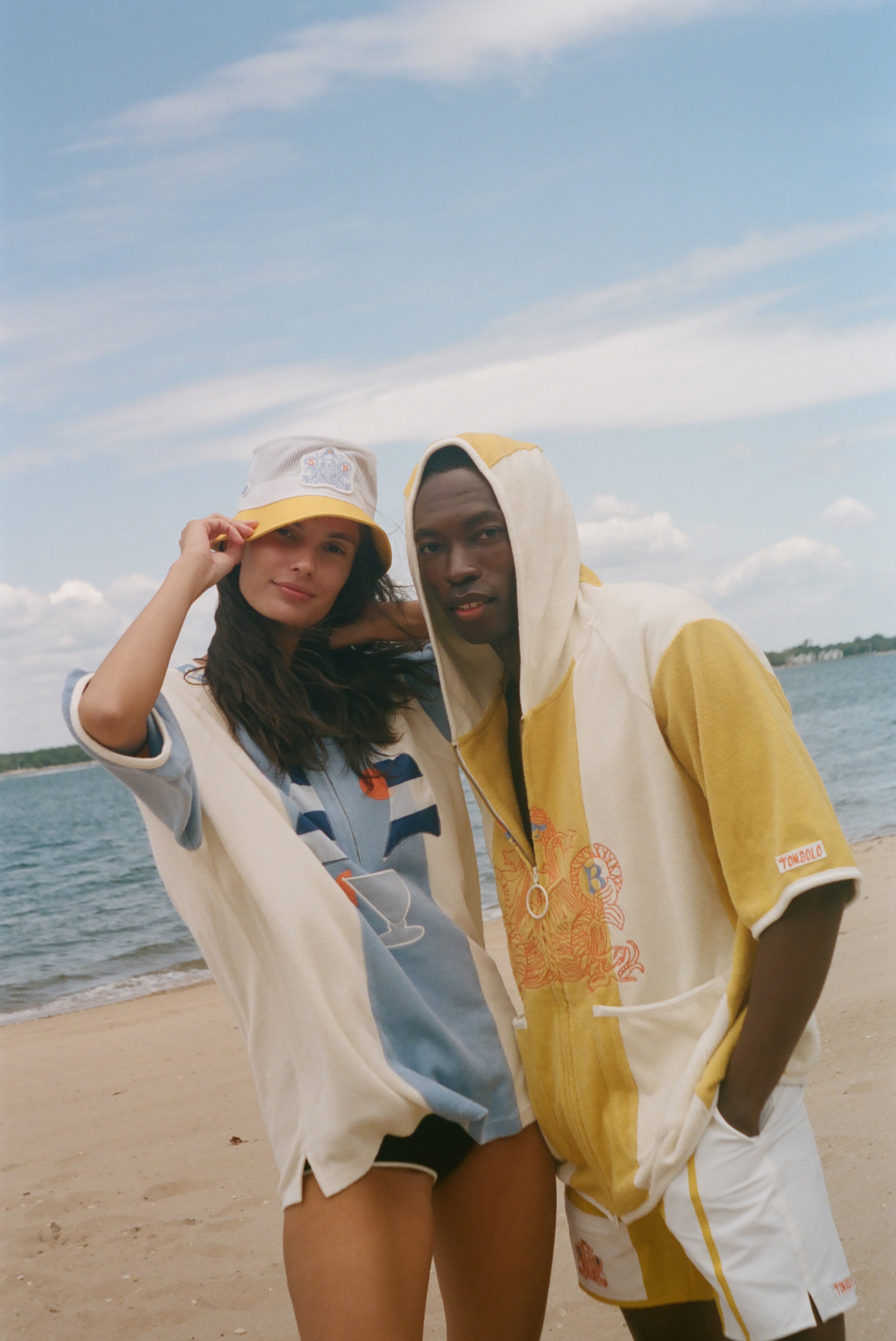 Two models standing on a sandy beach wearing matching yellow and off-white hooded beach poncho-style tops under a blue sky.
