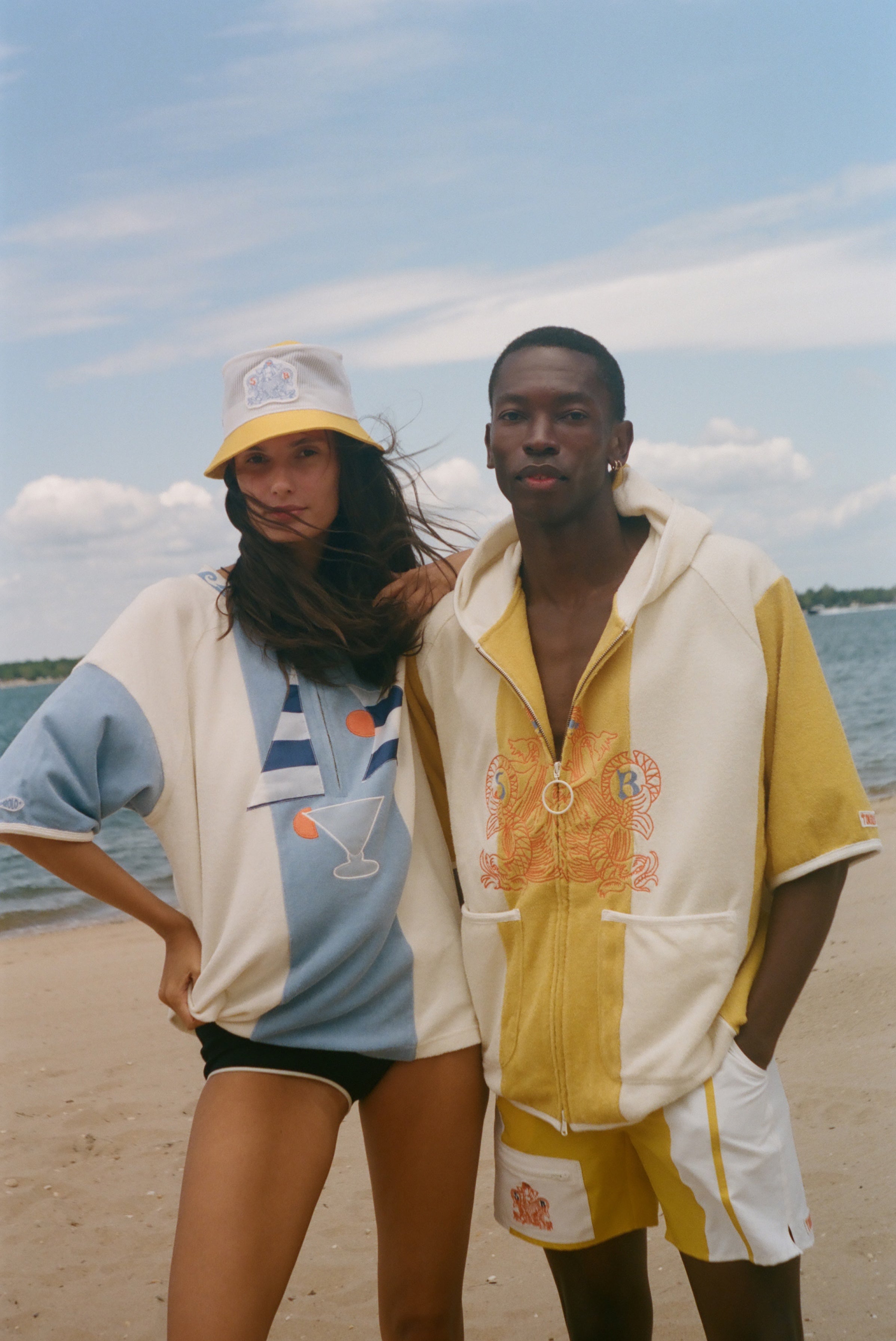 Two models posing on a sandy beach wearing matching color-blocked hoodies and shorts sets. The set features a color-blocked design of cream, yellow, and light blue, with a tropical cocktail graphic on the woman's hoodie and a stylized graphic on the man's hoodie.