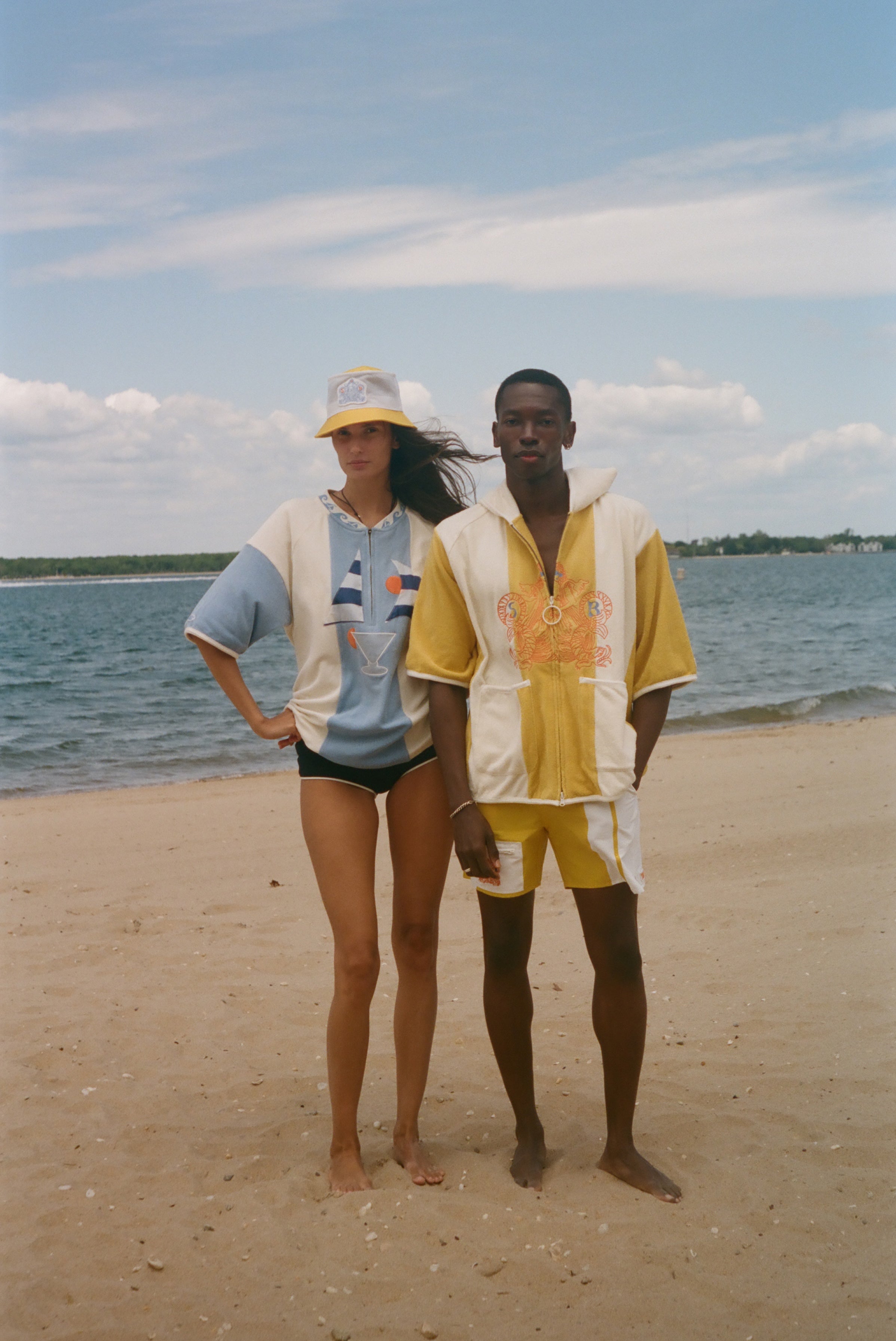 Two models standing on a sandy beach wearing matching two-piece swimwear sets. The woman wears a light blue and white set with a yellow and white cap, and the man wears a yellow and white set with a yellow and white cap.