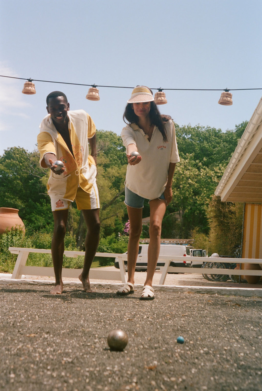 Two people outdoors on a gravel surface under a blue sky, wearing matching cream and yellow lounge sets while playing with small balls. A house, trees, and hanging string lights are visible in the background.