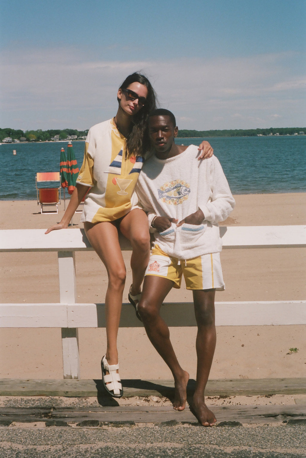 A couple posing in matching yellow and white swimwear and loungewear sets on a beach boardwalk with a white fence, ocean, and sky in the background.
