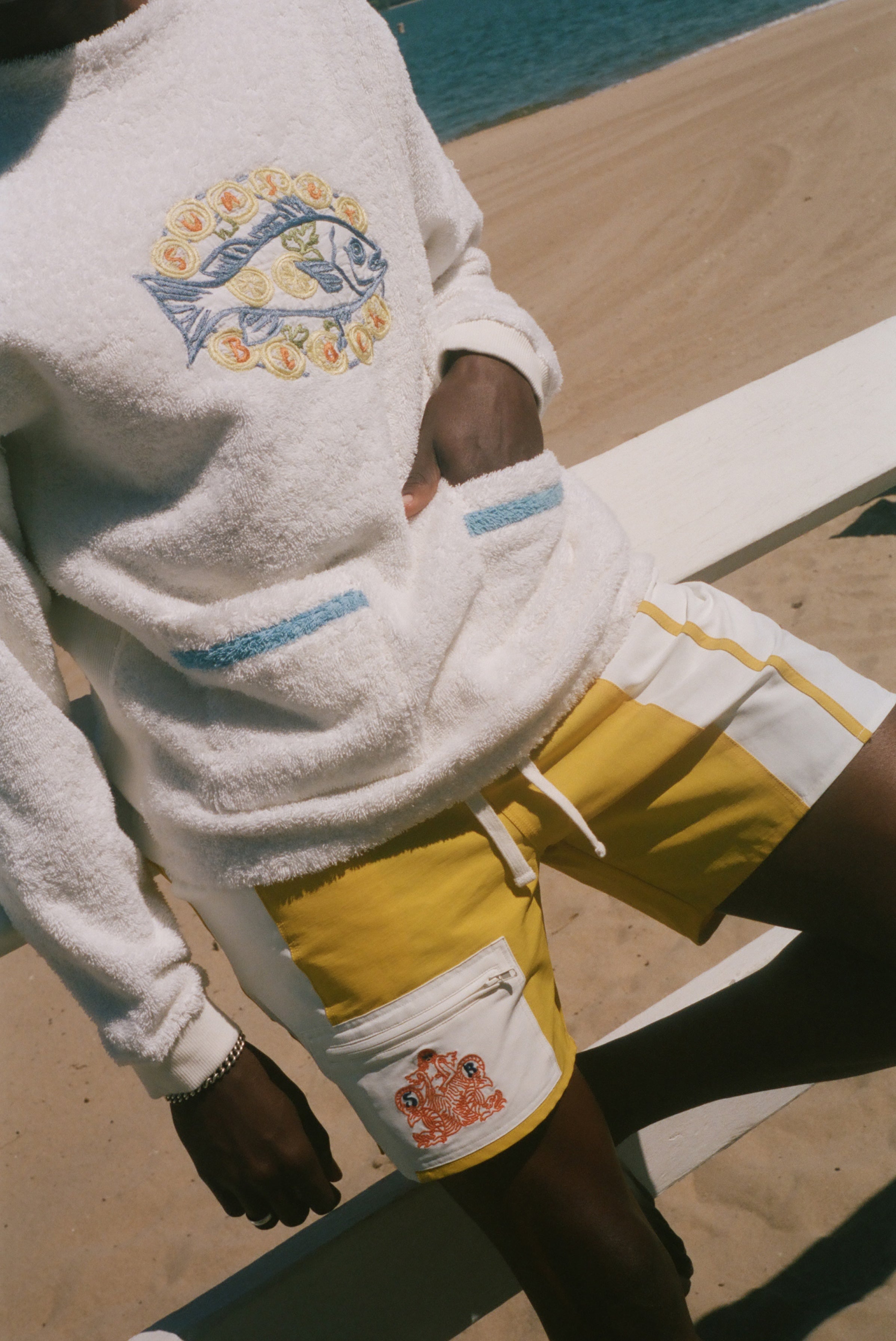 A person at a beach wearing a white terry sweatshirt with a blue and yellow fish graphic on the chest, white shorts with yellow side panels and a white front pocket featuring a small orange graphic, and dark swim trunks.