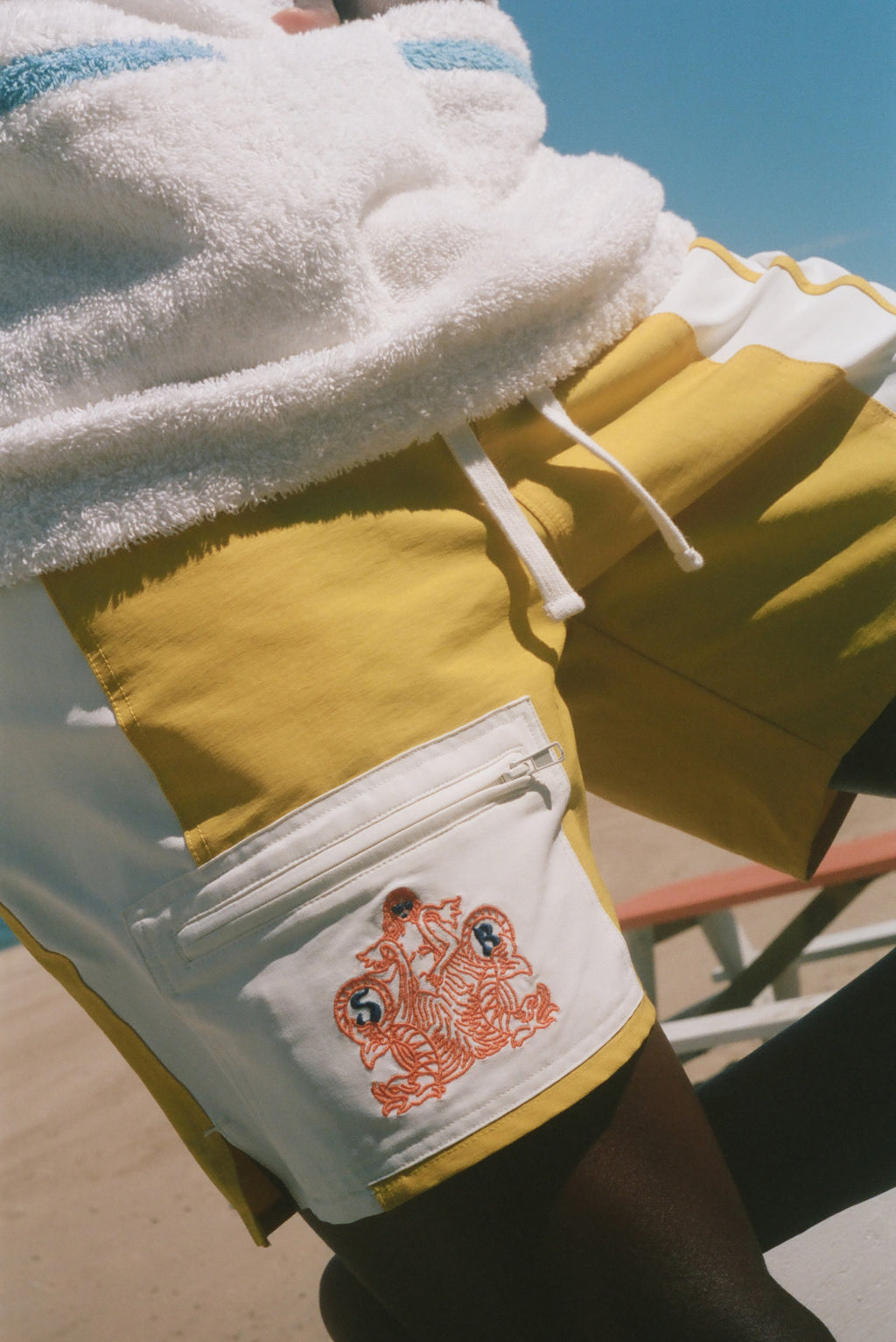 Close-up of a person wearing yellow and white board shorts with a white fleece-lined pocket featuring a red graphic print, under a white towel, with a blue sky background.