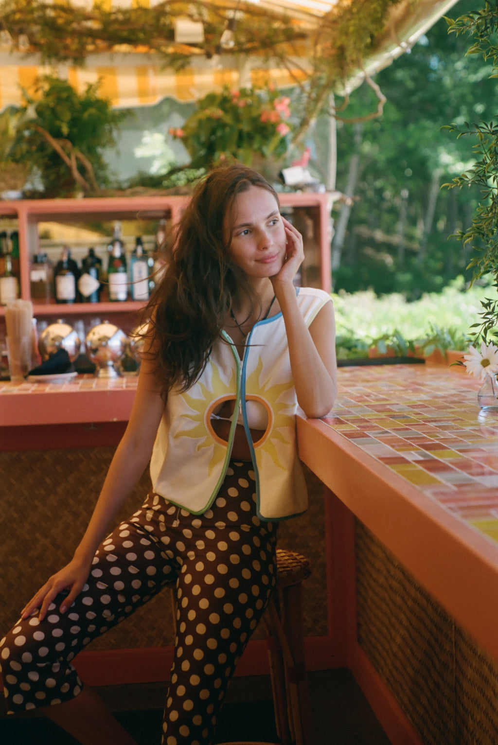A woman posing in a vibrant, tropical-themed bar or cafe, wearing a yellow sun graphic vest over a white top and black pants with white polka dots.