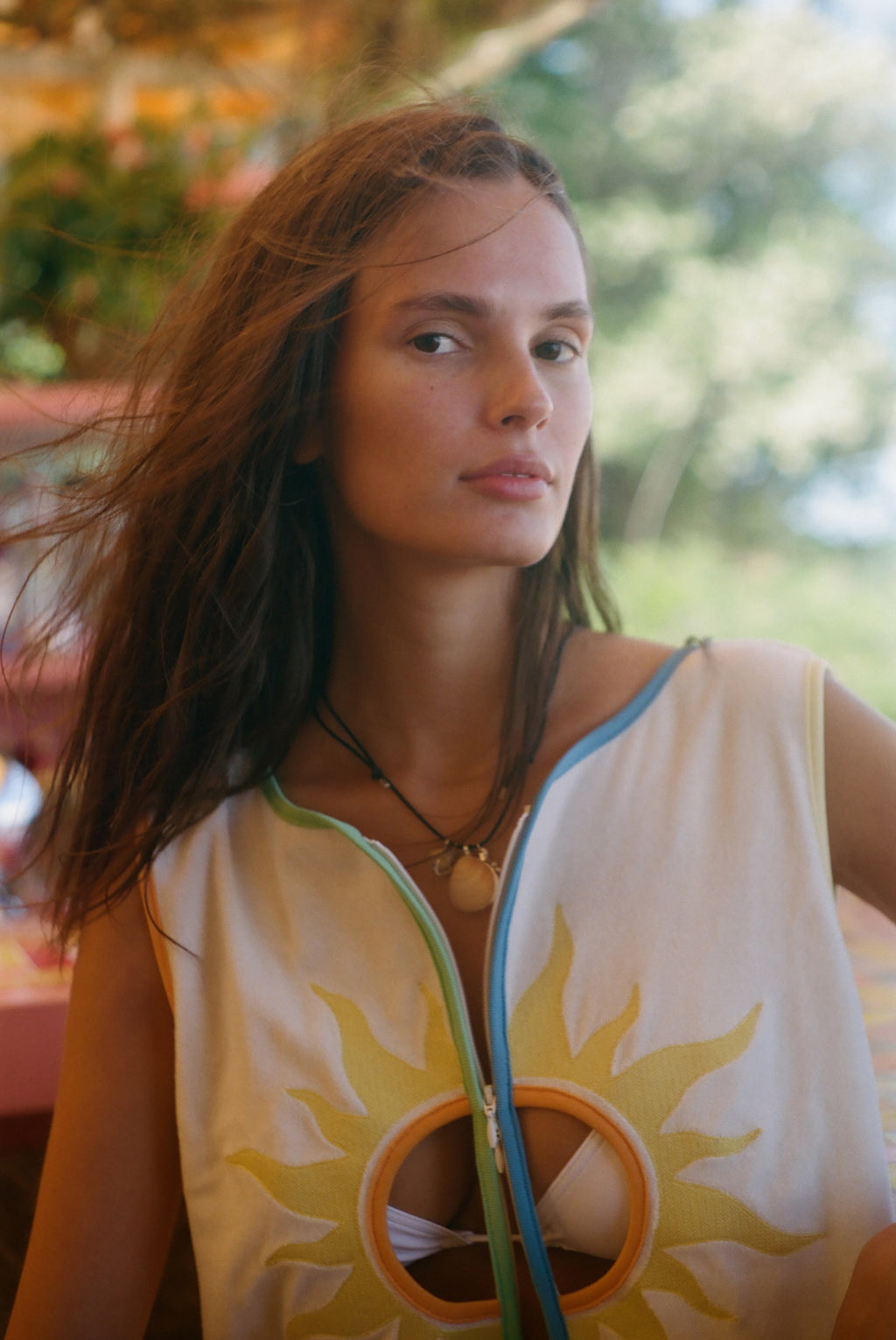 A woman wearing a sleeveless white top with a yellow sun graphic and colorful trim, photographed outdoors with a blurred green background.