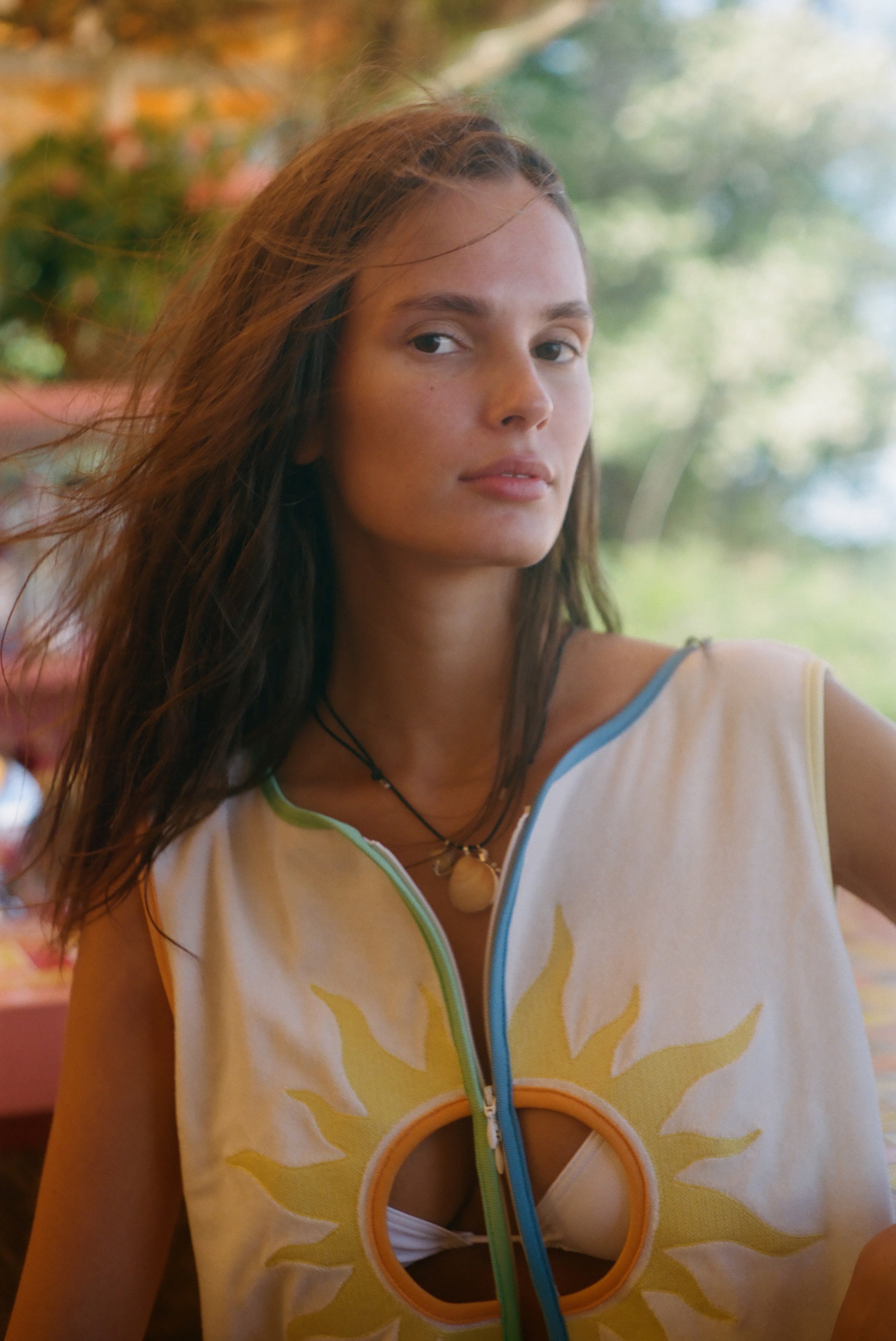 A woman wearing a sleeveless white top with a yellow sun graphic and colorful trim, photographed outdoors with a blurred green background.