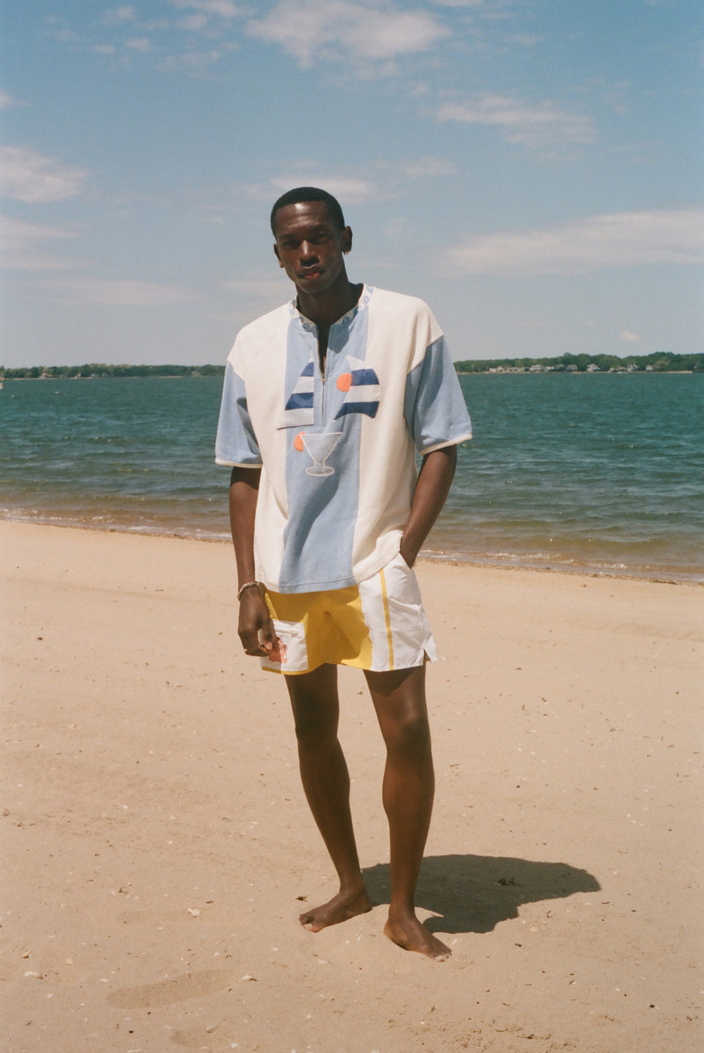 A man stands on a sandy beach wearing a color-blocked short-sleeve polo shirt and yellow-and-white shorts, with the ocean and blue sky in the background.