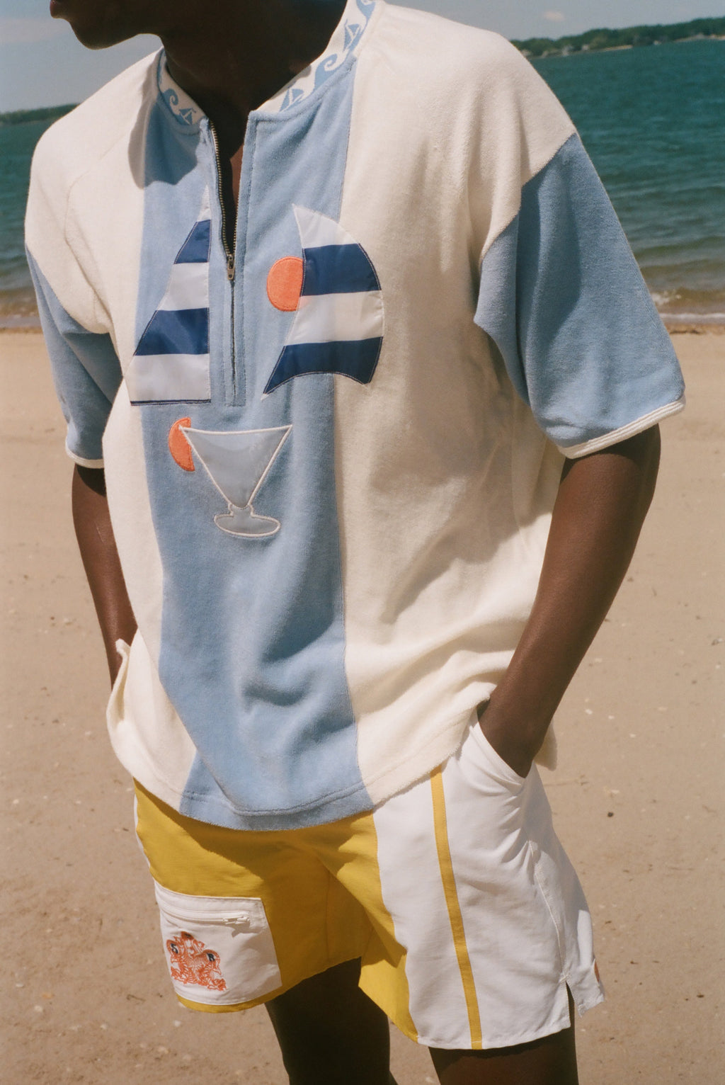 A man wearing a light blue and off-white color-blocked short-sleeve polo shirt with a half-zip collar, featuring a graphic print of a sailboat and a cocktail glass on the front. He is also wearing yellow and white shorts, standing on a sandy beach with the ocean in the background.