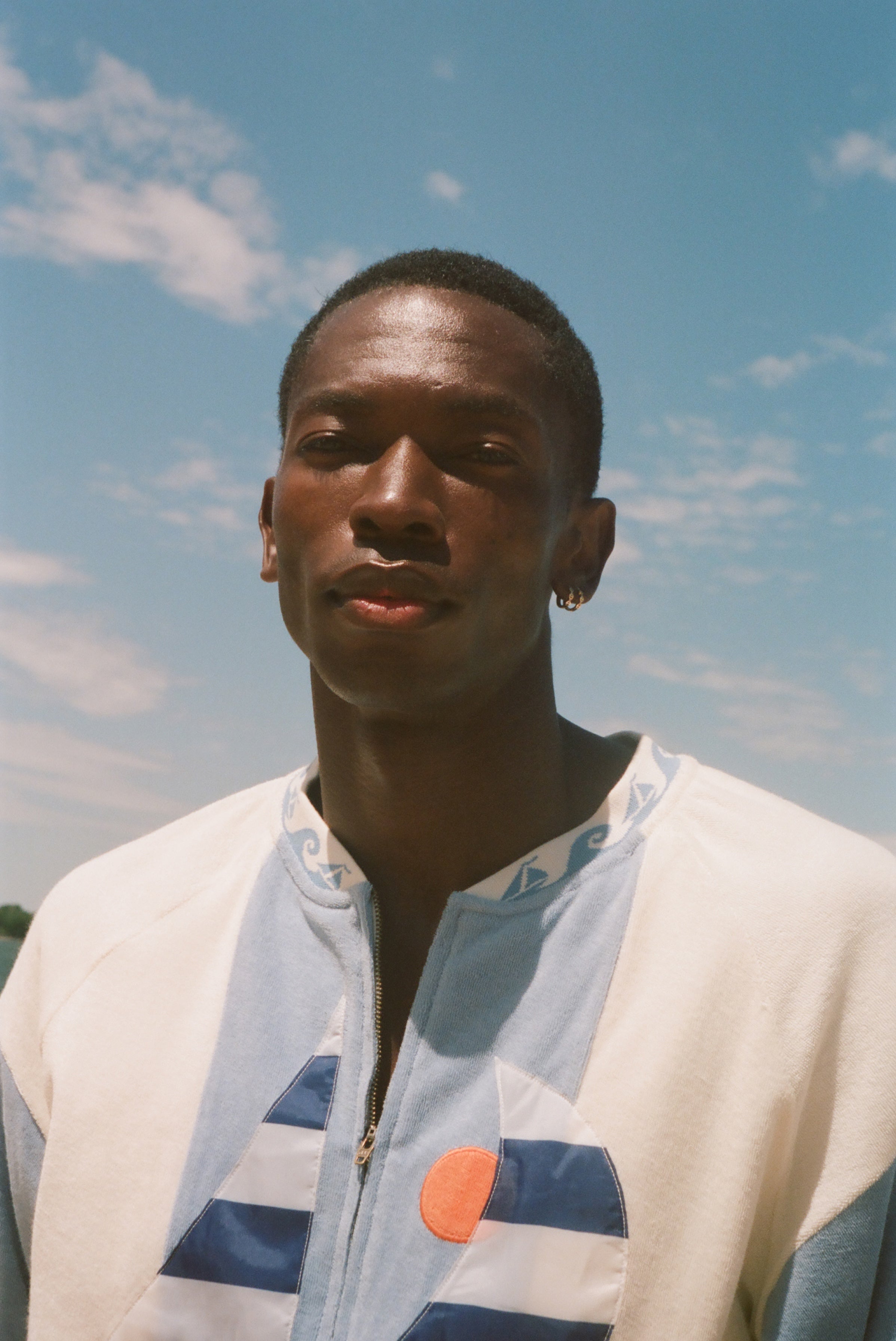 A man wearing a white zip-up shirt with blue and orange graphic details, photographed outdoors against a blue sky with light clouds.