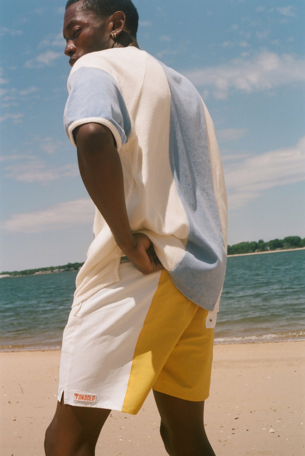 A man standing on a sandy beach wearing a color-blocked short-sleeve cabana shirt. The shirt features cream, light blue, and gray color blocking, while the shorts are white with a yellow side panel.
