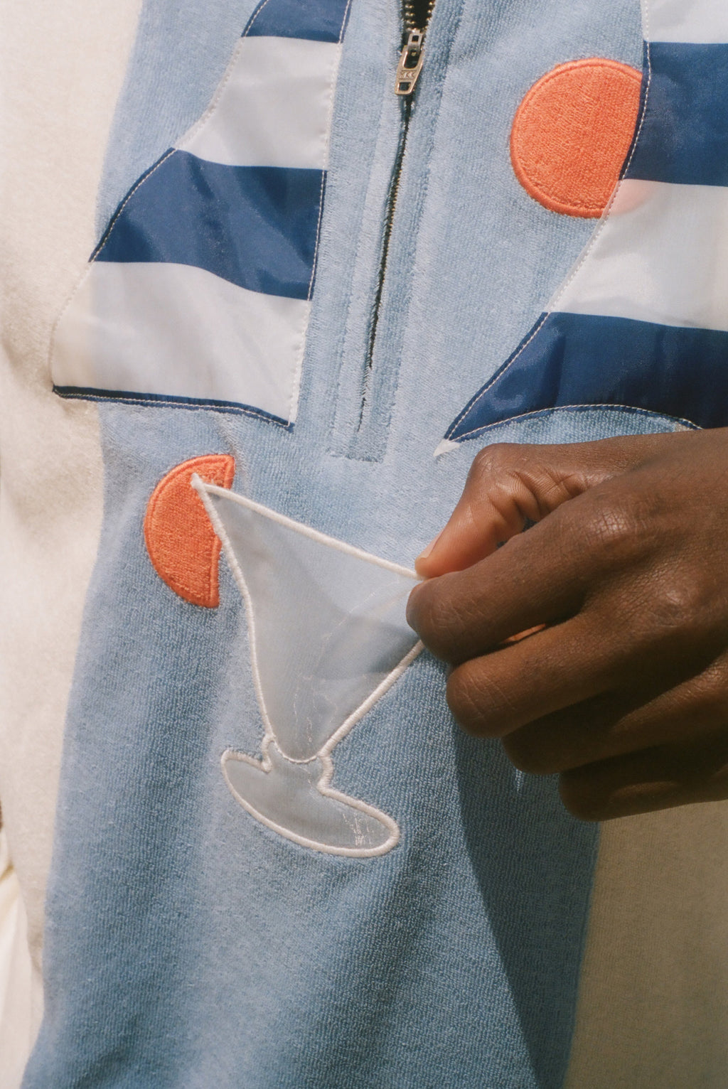 A close-up of a person wearing a light blue zip-up jacket with navy and white stripes, orange circular patches, and a white embroidered cocktail glass motif, while holding a small white fabric piece.