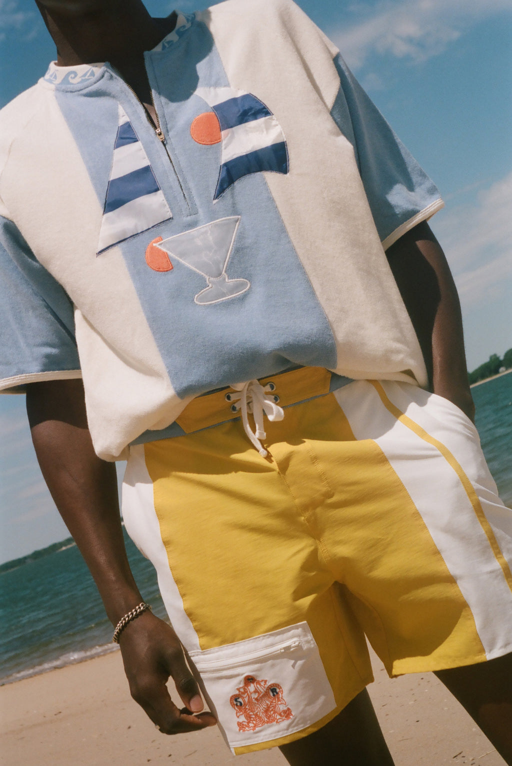 A person standing on a beach wearing a light blue and cream short-sleeve polo shirt with embroidered sailboats and a martini glass, paired with yellow and white drawstring shorts with a white cargo pocket.