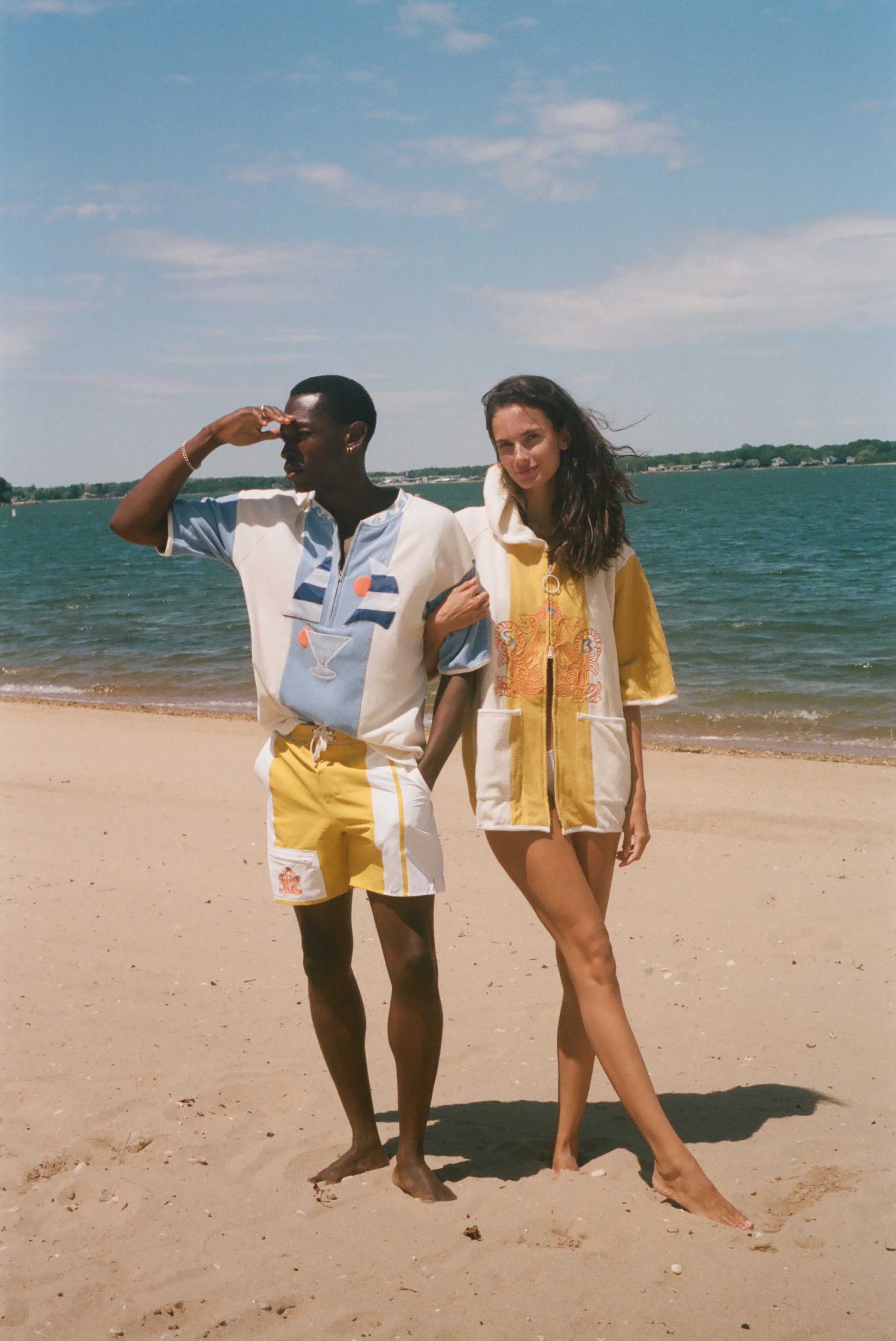 A man and a woman standing on a sandy beach wearing matching vintage-style swimwear sets. The man wears a white and light blue short-sleeve collared swim shirt with yellow and white shorts, while the woman wears a yellow and white patterned short-sleeve cover-up shirt with matching yellow and white bottoms.