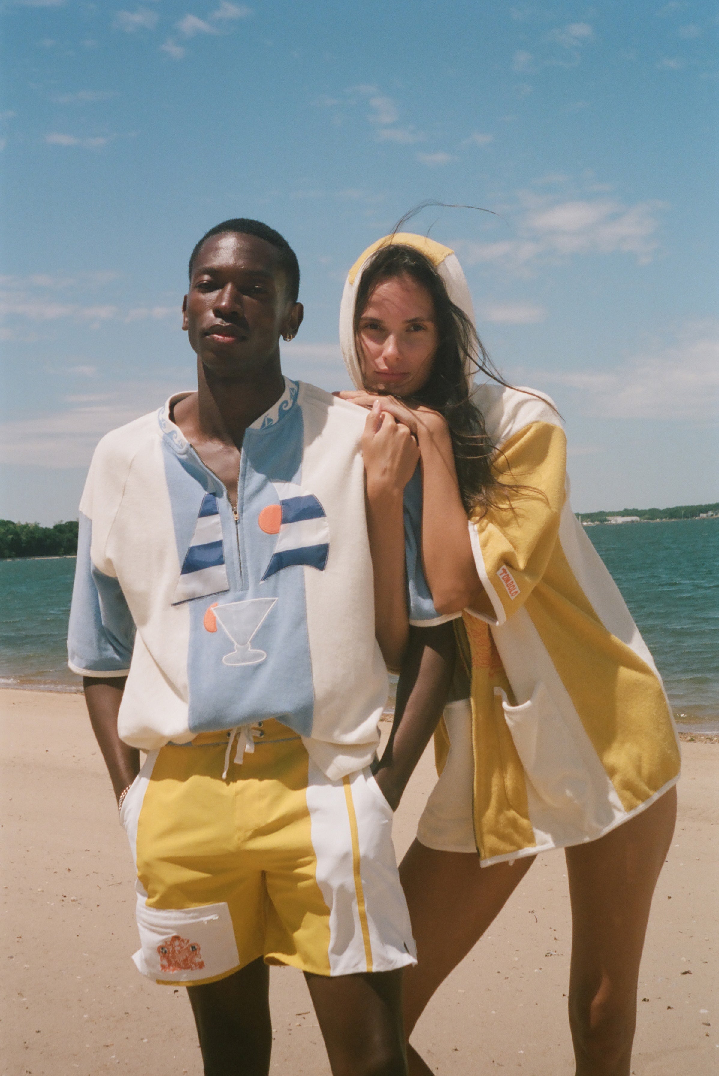A man and a woman standing on a sandy beach wearing matching light blue and yellow resort wear, with the ocean and blue sky in the background.