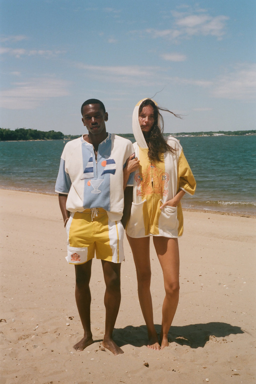 Two models standing on a sandy beach wearing matching yellow and off-white hooded beach poncho-style tops under a blue sky.