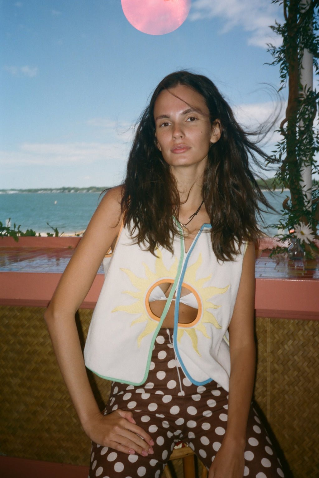 A woman posing outdoors by the water wearing a sleeveless white vest with a yellow sun graphic and brown polka-dot pants.