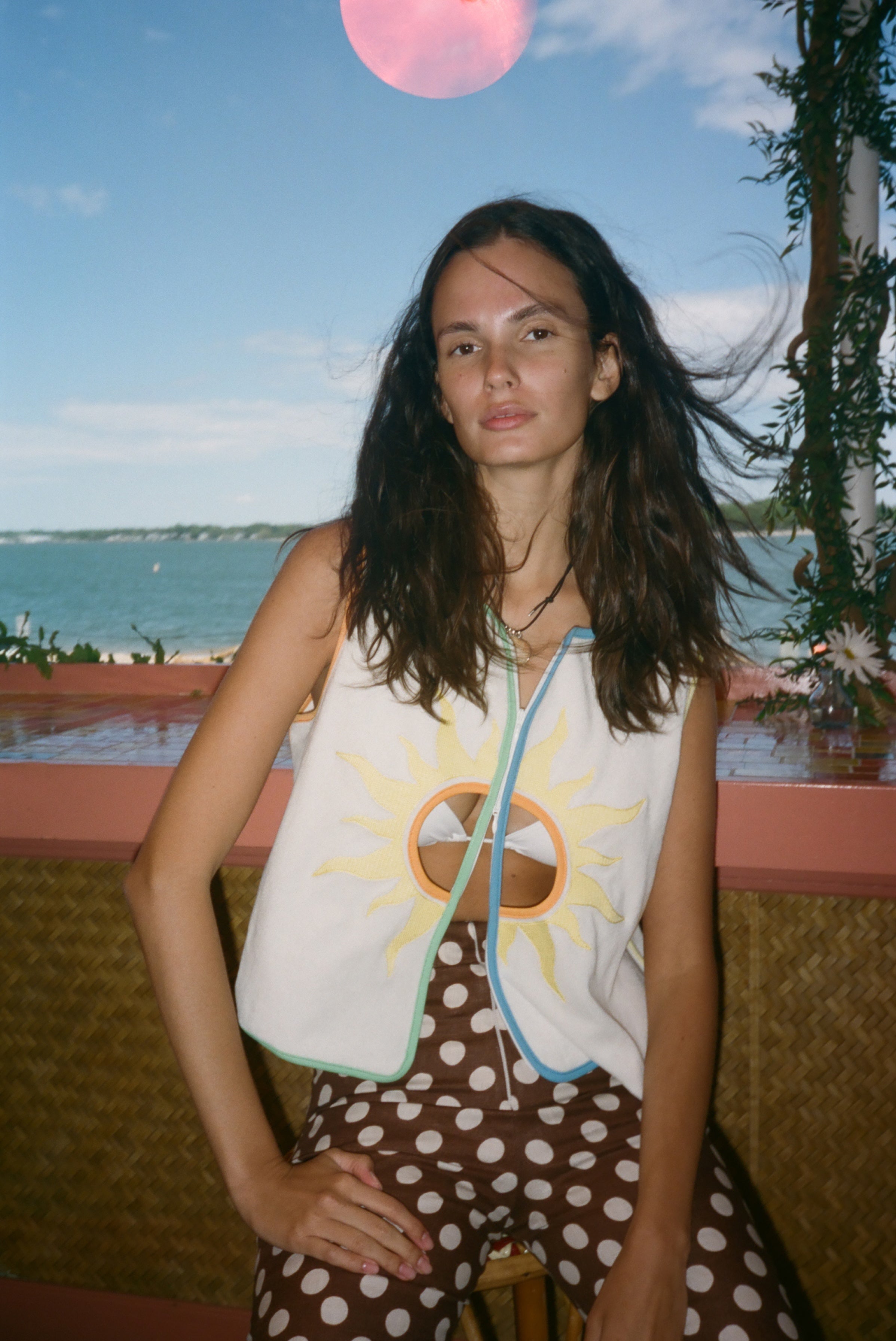 A woman posing outdoors by the water wearing a sleeveless white vest with a yellow sun graphic and brown polka-dot pants.