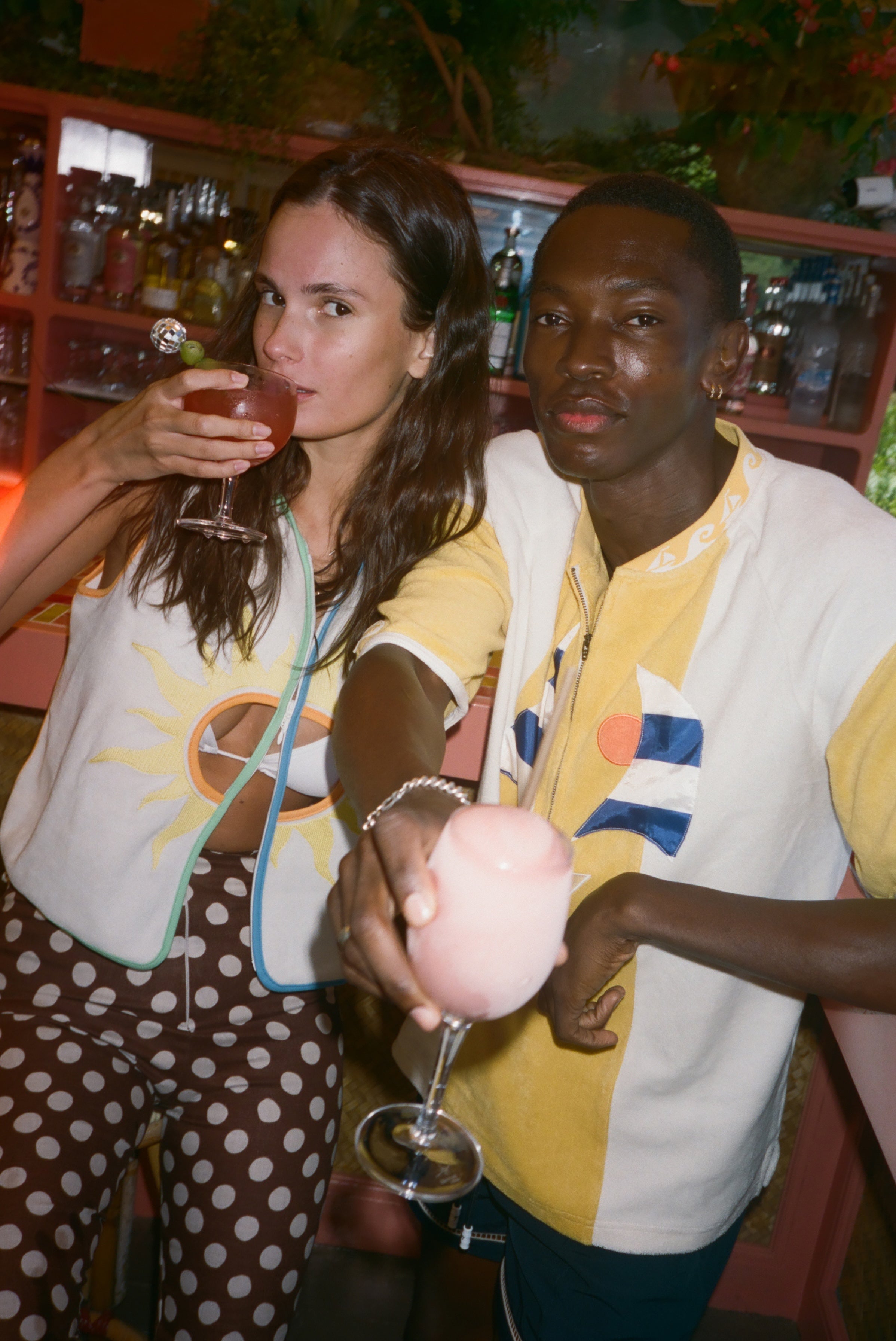 A man and a woman in a dimly lit bar setting, both wearing Sunset Beach collection items.