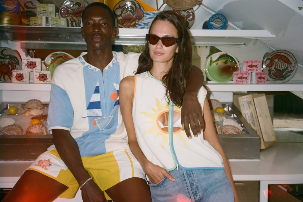 Two models posing together in a shop setting. The man wears a light blue and white striped polo shirt with yellow shorts, and the woman wears a white sleeveless top with a yellow sun graphic, denim shorts, and sunglasses.