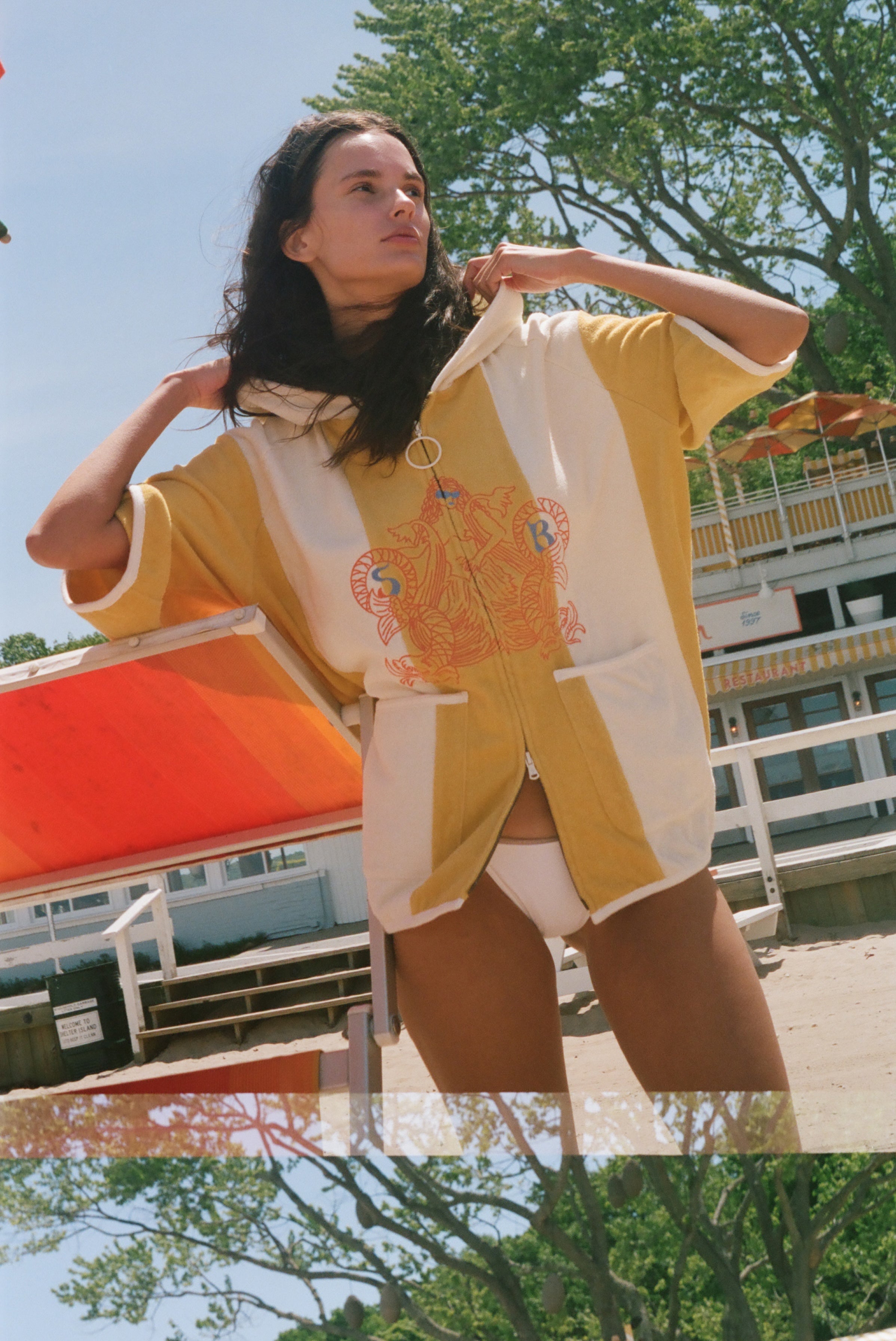 A woman wearing a yellow and white oversized beach poncho with colorful embroidery on the front, standing outdoors near a beach chair and trees.