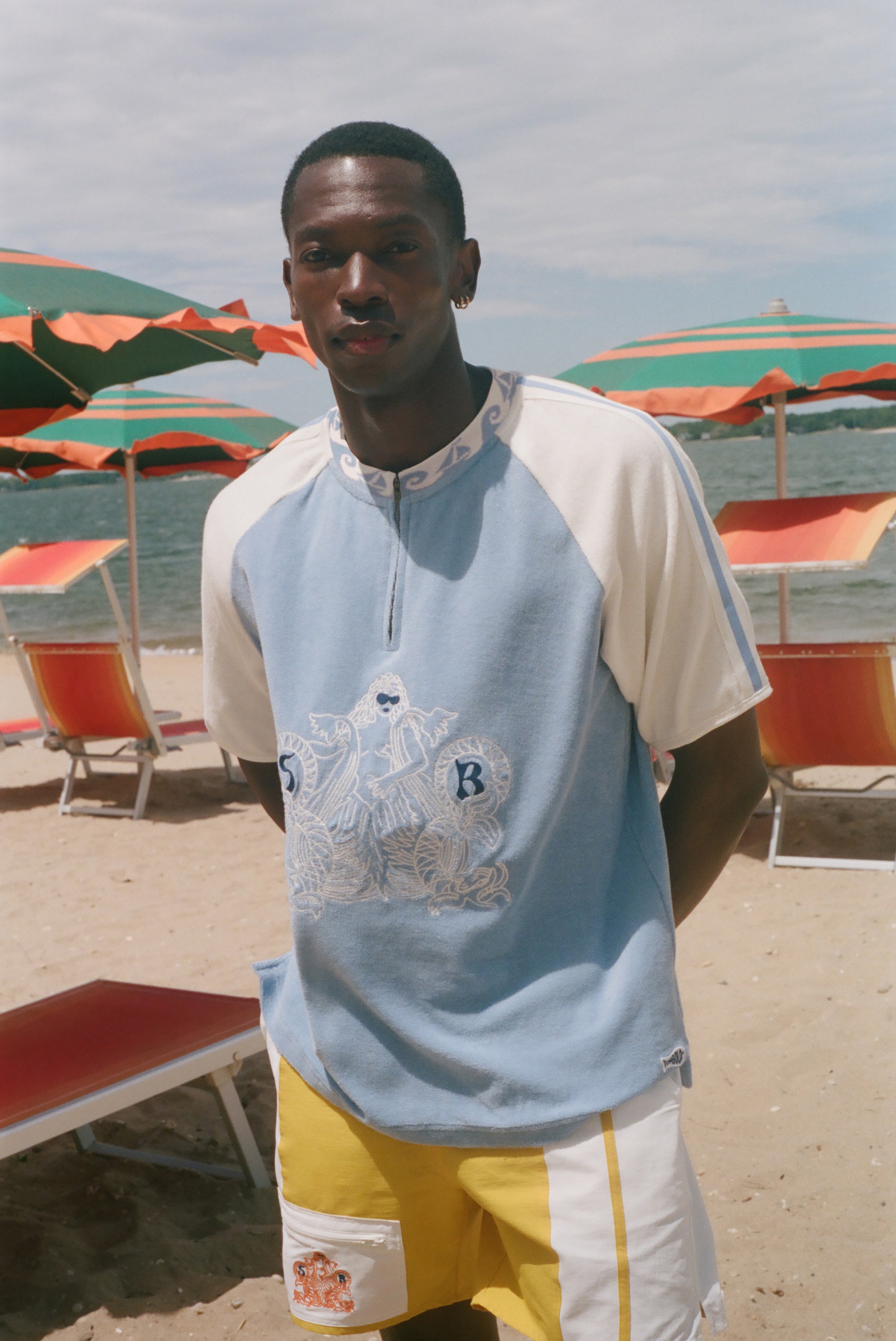 A man standing on a sandy beach wearing a light blue and off-white raglan t-shirt with a graphic print on the front, paired with yellow and white shorts. Beach umbrellas and lounge chairs are visible in the background.