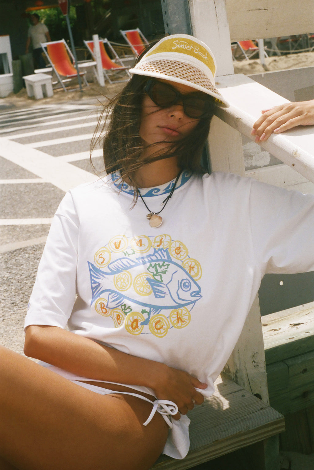 A woman wearing a white graphic t-shirt with a blue and yellow fish design, a straw hat, and sunglasses, sitting outdoors on a wooden bench.