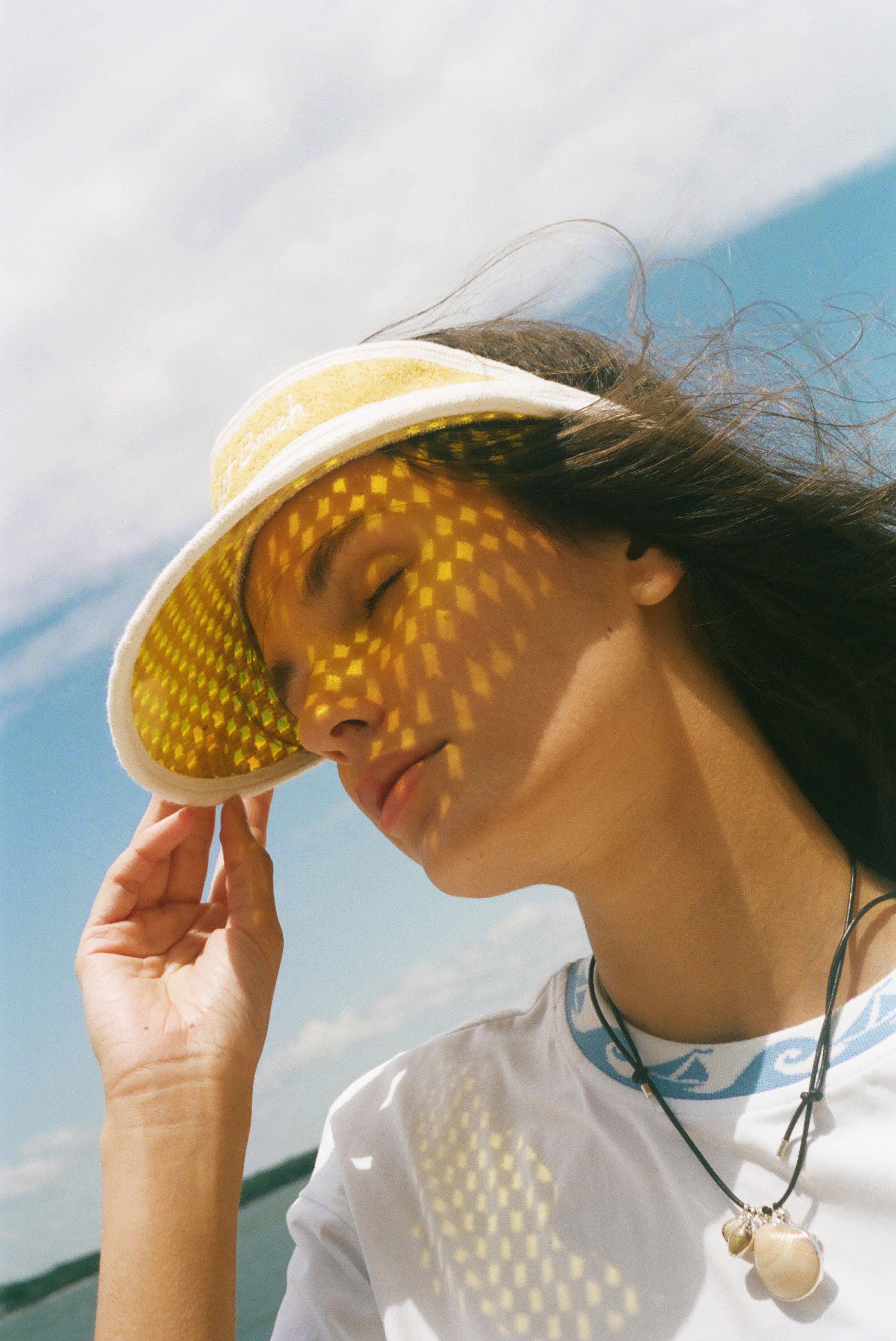 A woman outdoors wearing a yellow sun visor with a geometric dot pattern, with sunlight casting a matching pattern of shadows across her face. She is wearing a white t-shirt and a necklace.