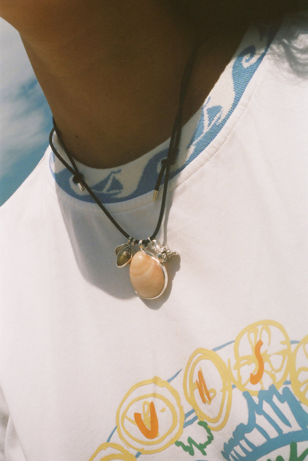 A close-up of a person wearing a dark cord necklace with a peach-colored shell pendant and small silver-toned accents, paired with a white graphic t-shirt.
