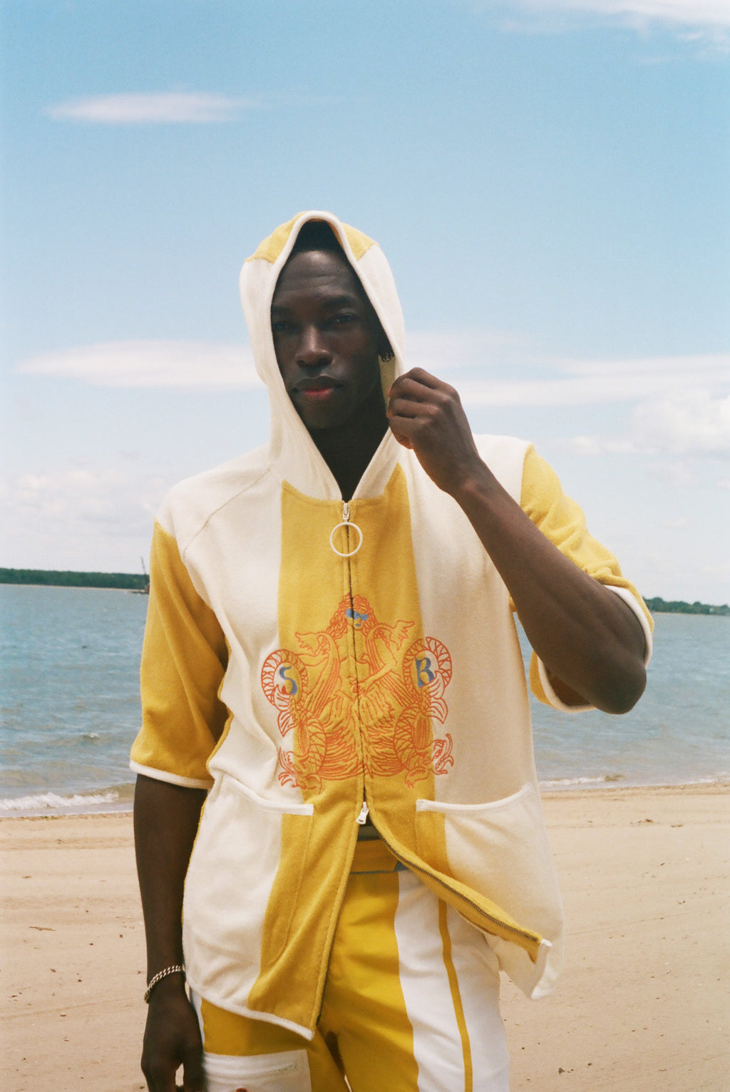 A man wearing a yellow and off-white hooded zip-up jacket with a large orange graphic on the front, standing on a sandy beach with water and sky in the background.