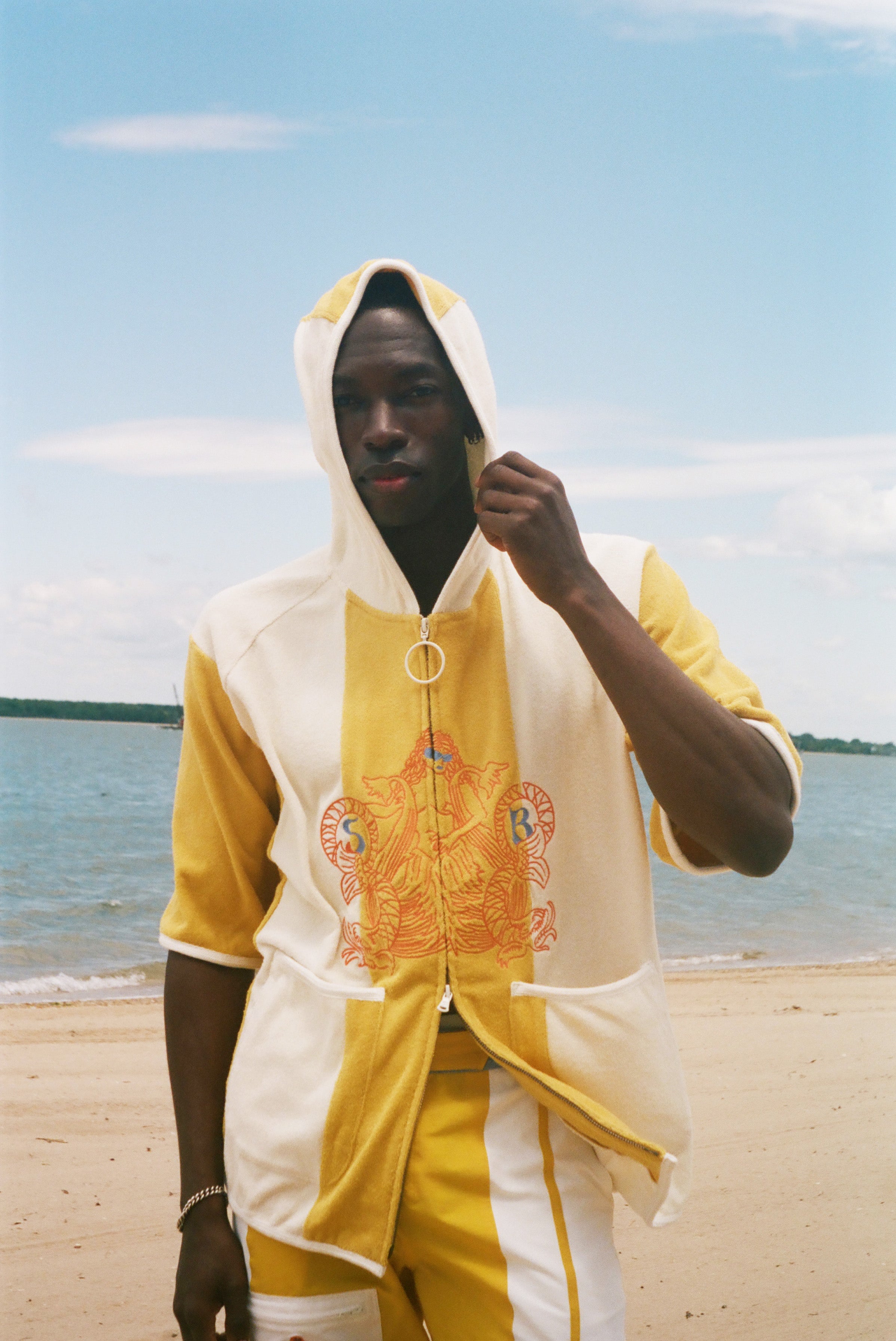 A man wearing a yellow and off-white hooded zip-up jacket with a large orange graphic on the front, standing on a sandy beach with water and sky in the background.