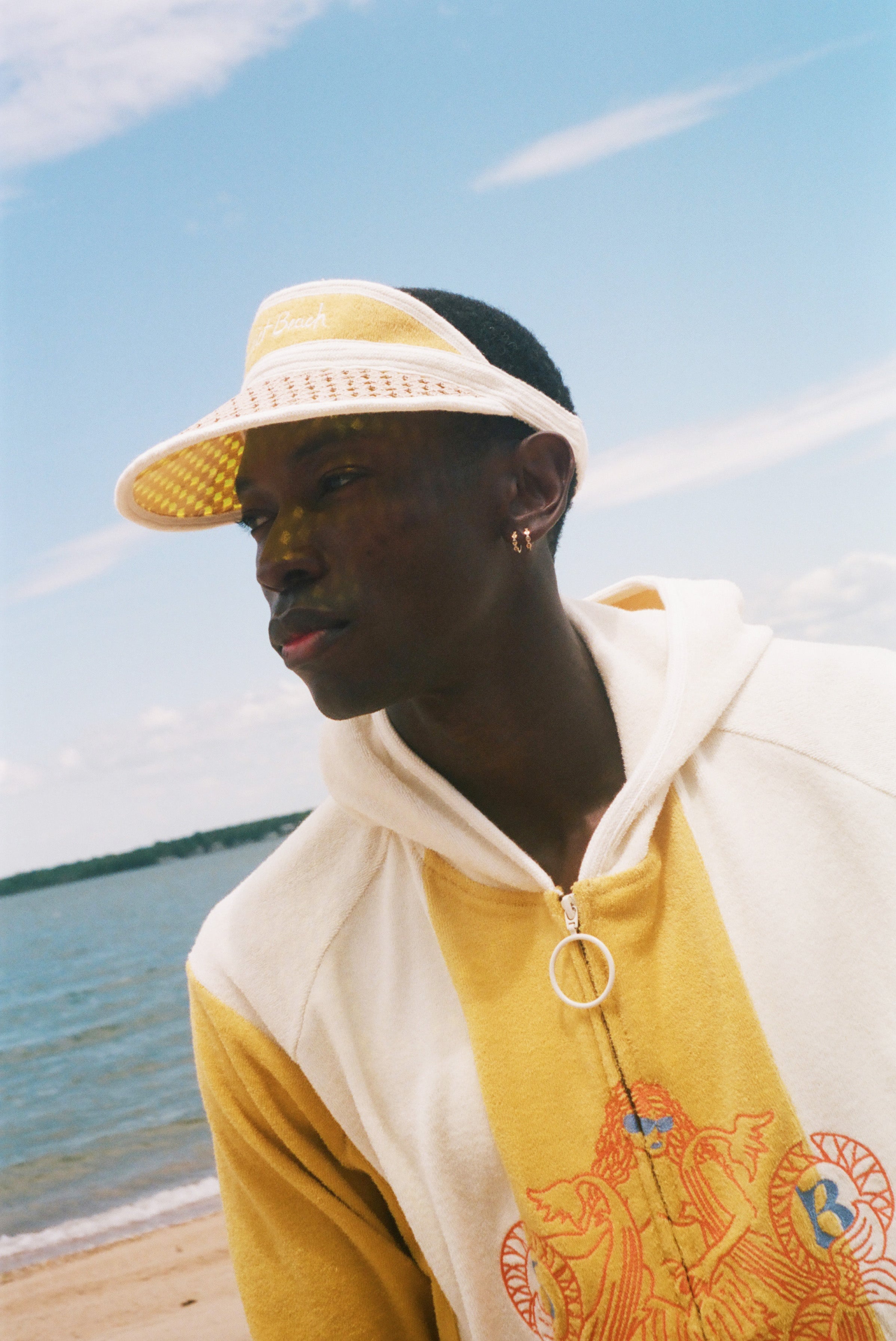 A man wearing a yellow and off-white hooded sweatshirt with a large graphic on the front, a matching yellow visor, and a black cap, standing on a beach with the ocean and blue sky in the background.