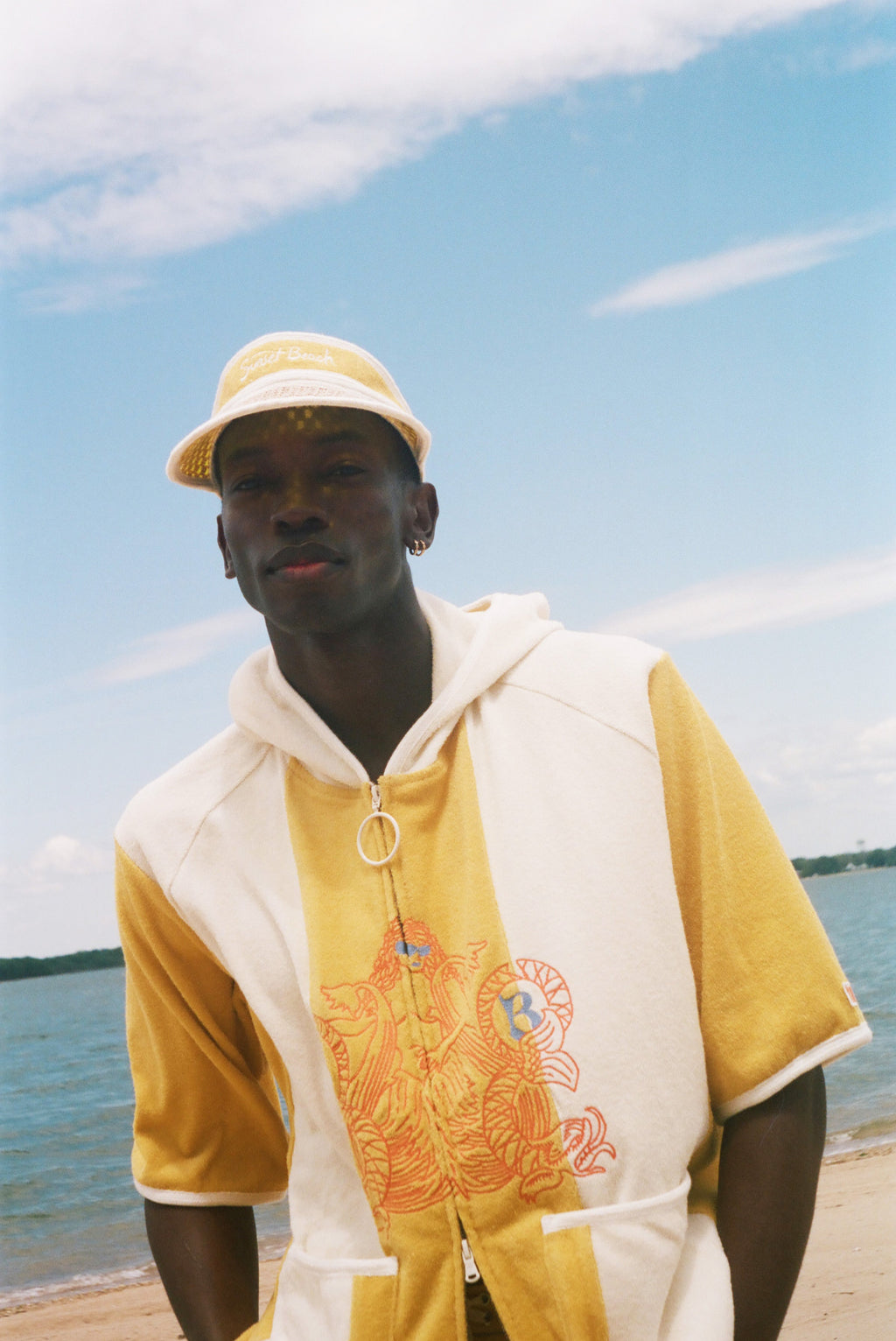 A man wearing a yellow and off-white hooded short-sleeve top with a graphic print stands on a beach with water and sky in the background.