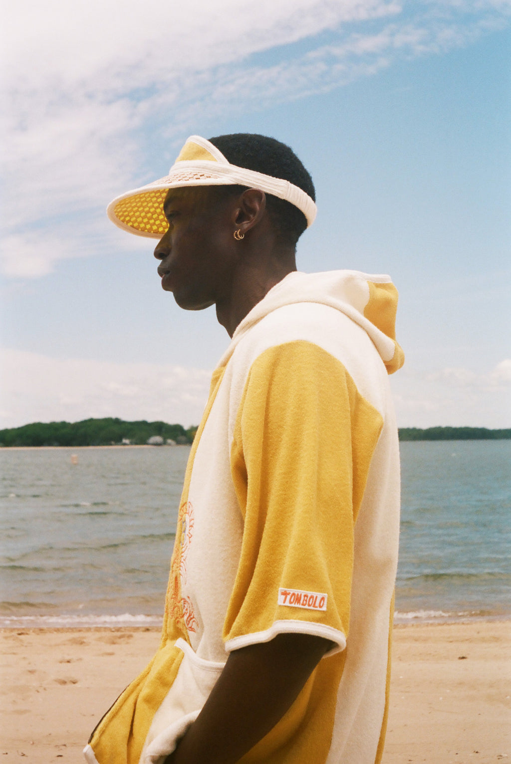 A man in profile wearing a yellow and off-white color-blocked hoodie and matching visor, standing on a sandy beach with water and sky in the background. 